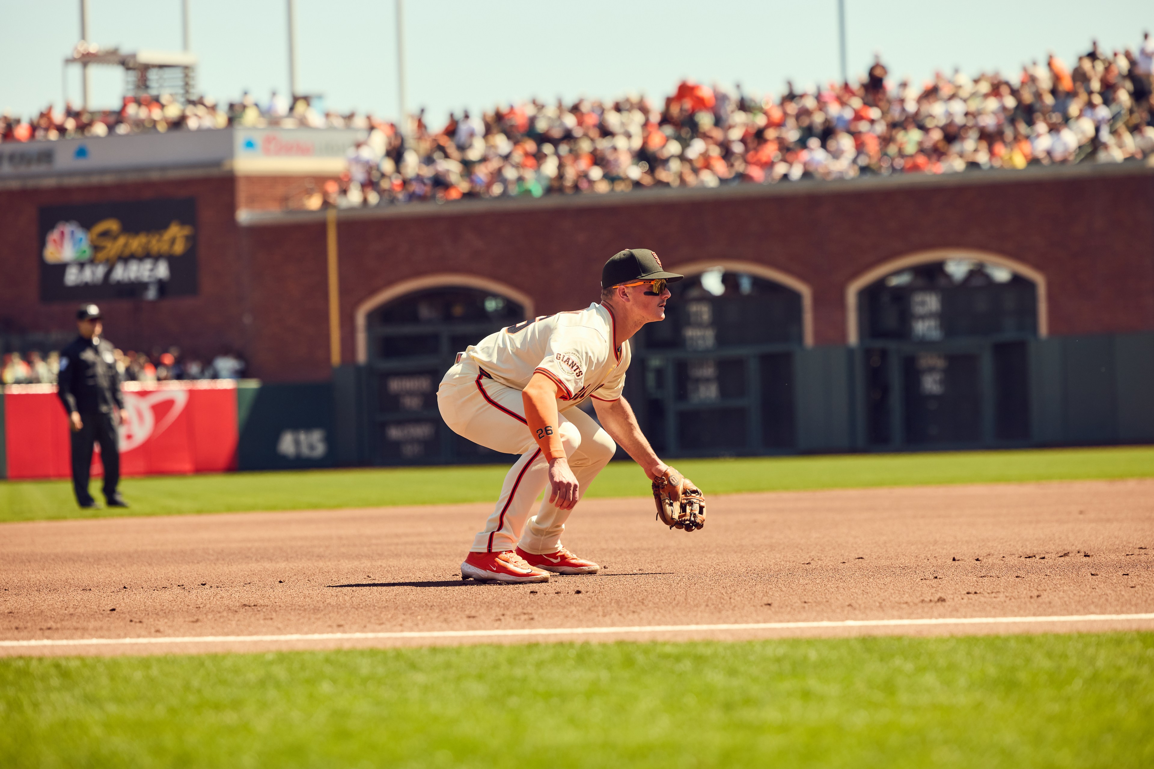 A baseball player in a white uniform is crouching in a field, poised and ready with a glove. A crowd watches from the stands in the background.