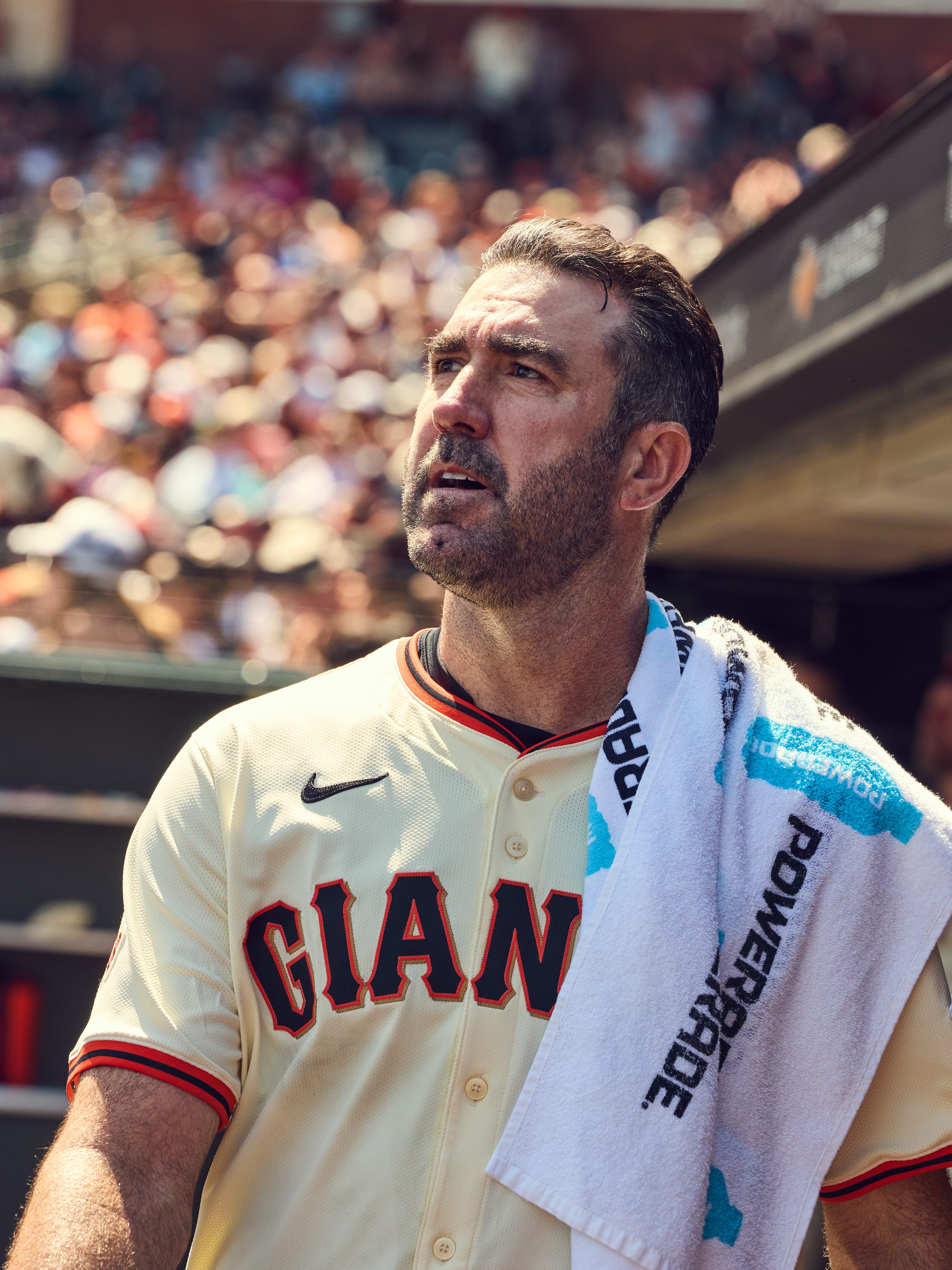 A baseball player wearing a &quot;GIANTS&quot; jersey looks upward, with a towel draped over his shoulder. A blurred crowd is visible in the background.