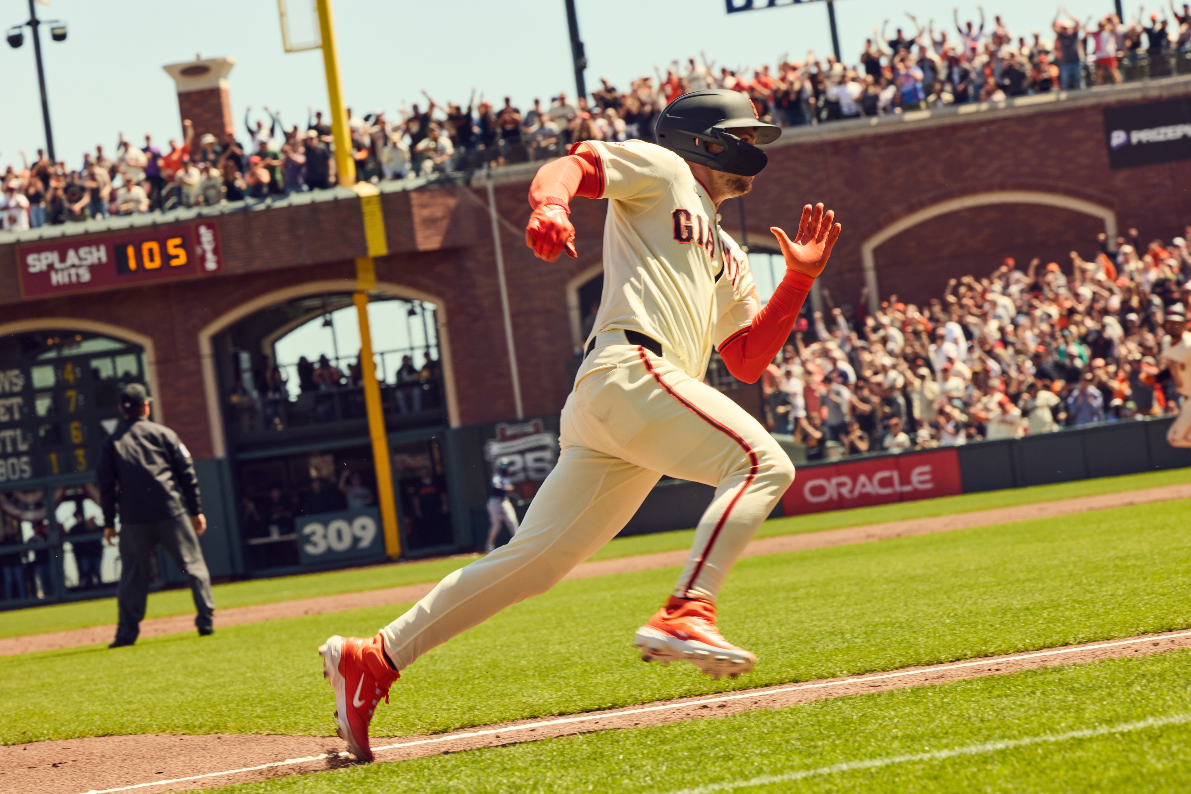 A baseball player runs bases on a field as a crowd in the background cheers, with a scoreboard visible. He wears a Giants uniform and bright orange cleats.