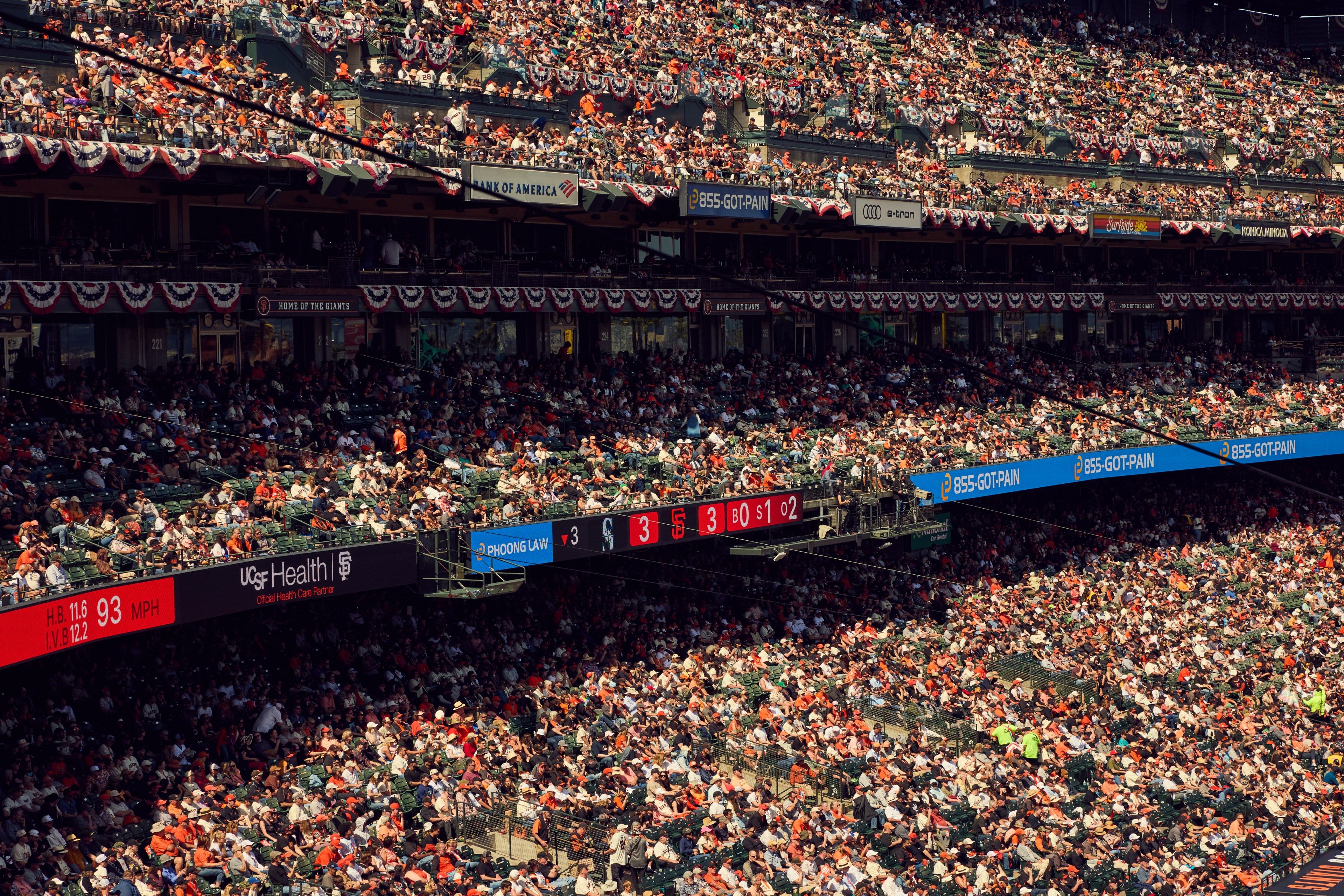 A packed baseball stadium with spectators filling the seats. Banners and ads are visible, along with a scoreboard indicating a tie game at 3-3.