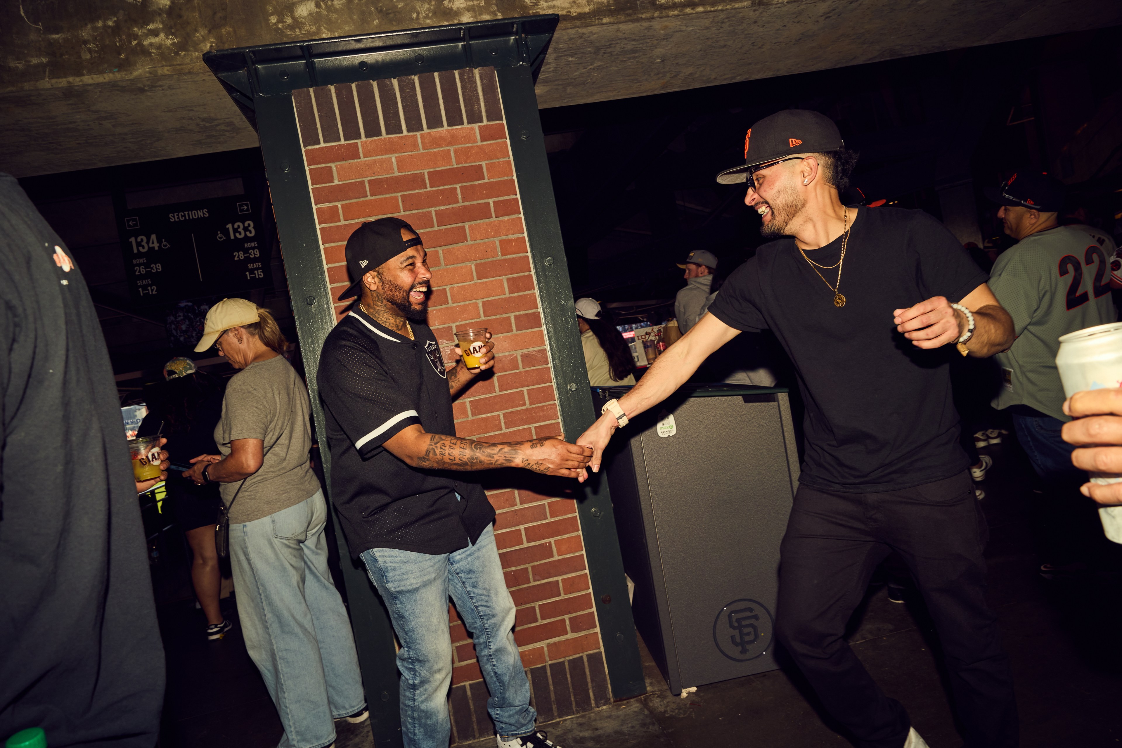 Two men, both wearing caps, joyfully shake hands near a brick column. One holds a drink, and they're surrounded by people in a lively setting.