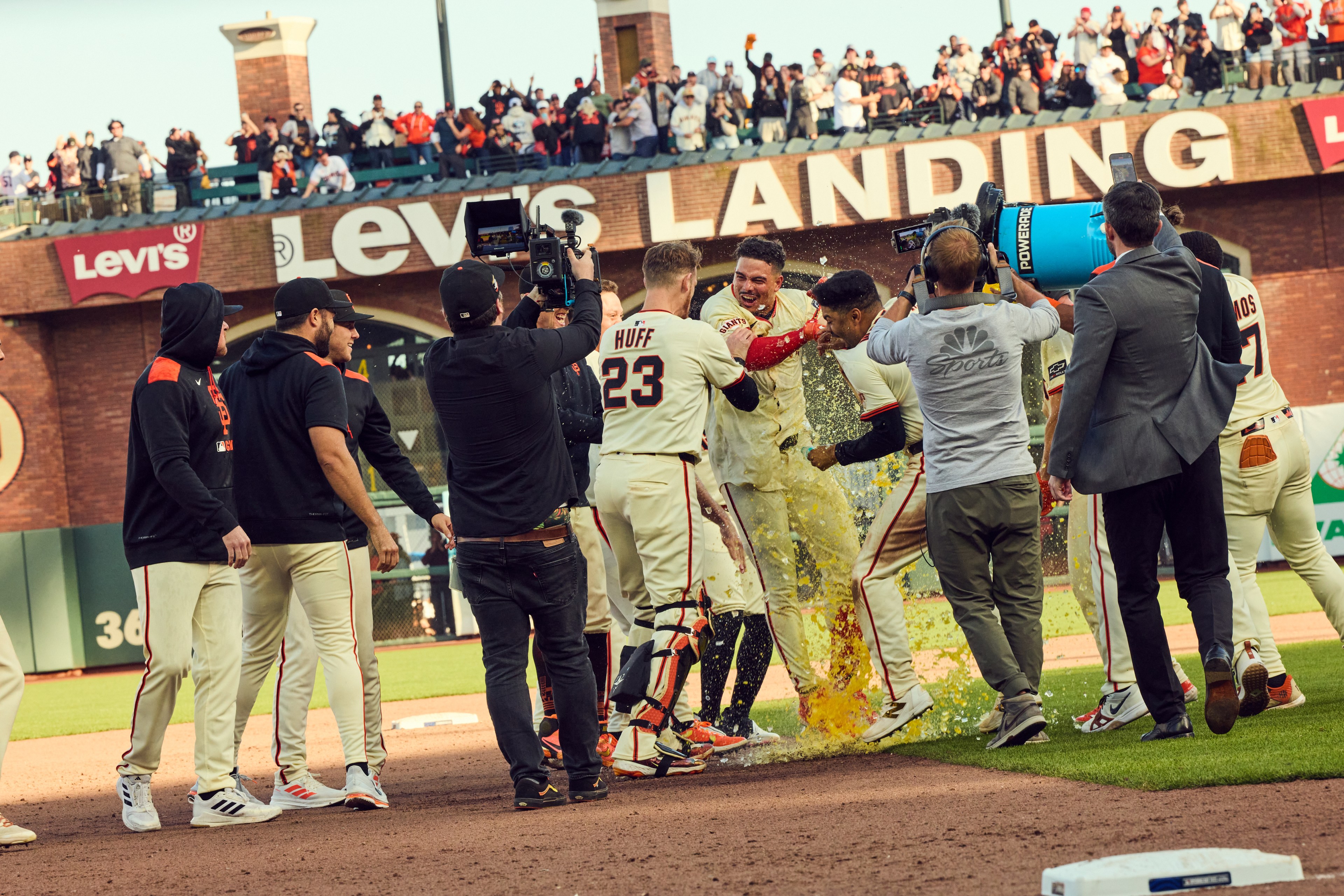 A group of baseball players celebrates on the field. One is doused with a cooler of liquid, while others cheer with excitement. Crowds watch from the stands.