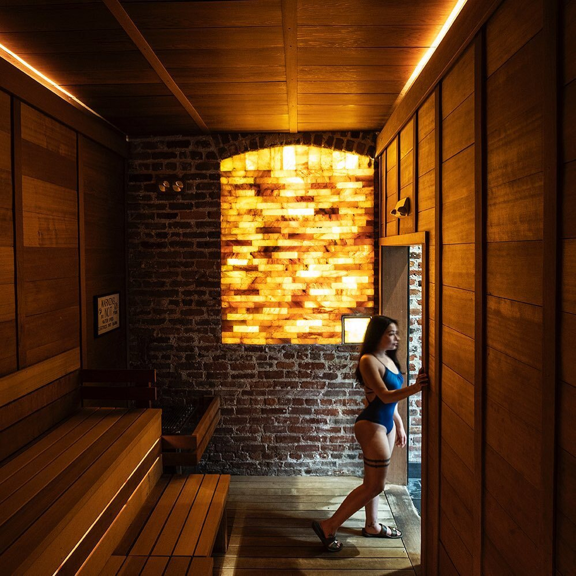 A woman in a swimsuit stands by the door of a cozy wooden sauna with glowing salt bricks, benches, and a sign on the brick wall.