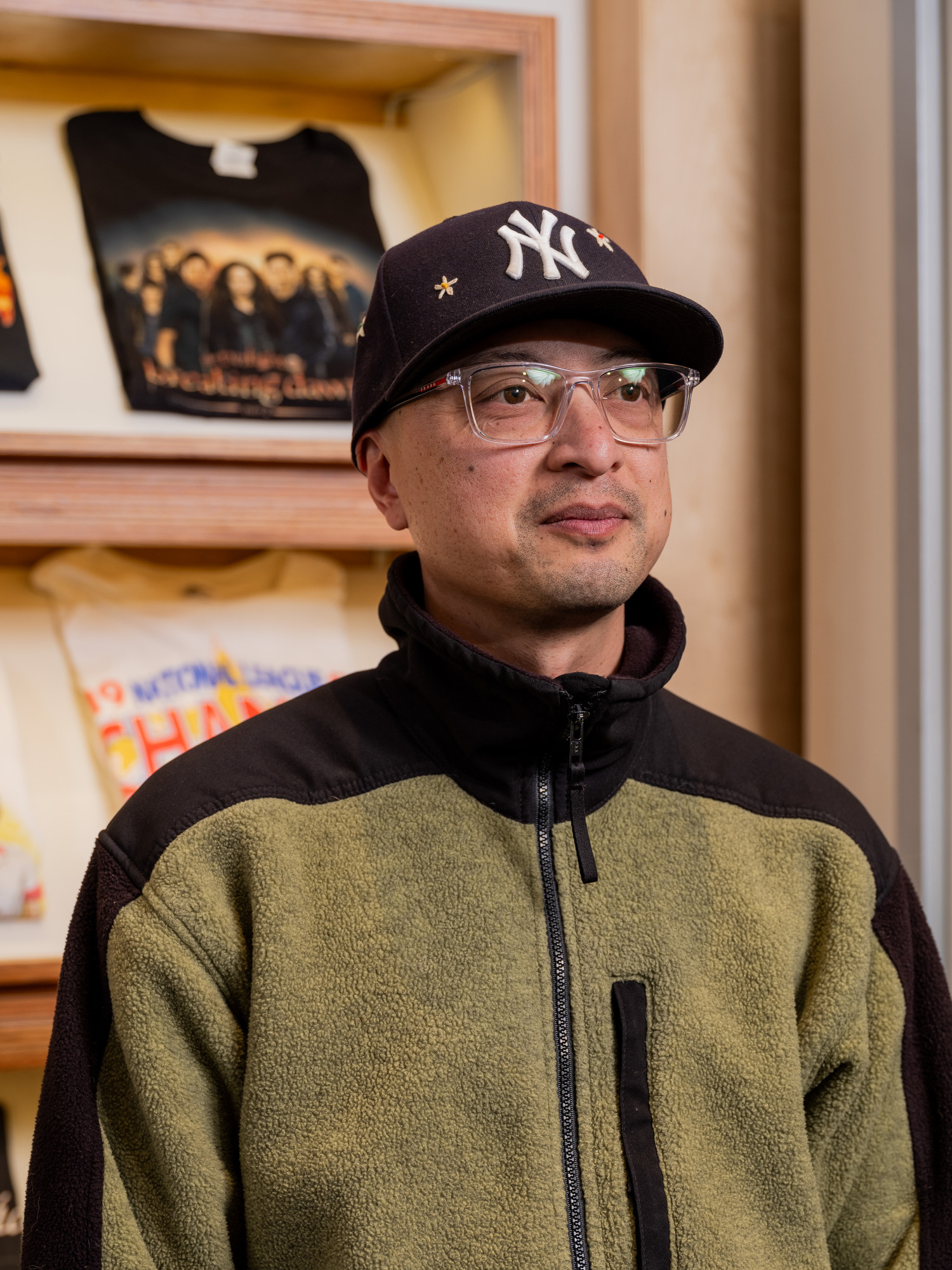 A person wearing glasses and a New York Yankees cap is in front of a wall display with folded t-shirts, including a black one featuring a group image.