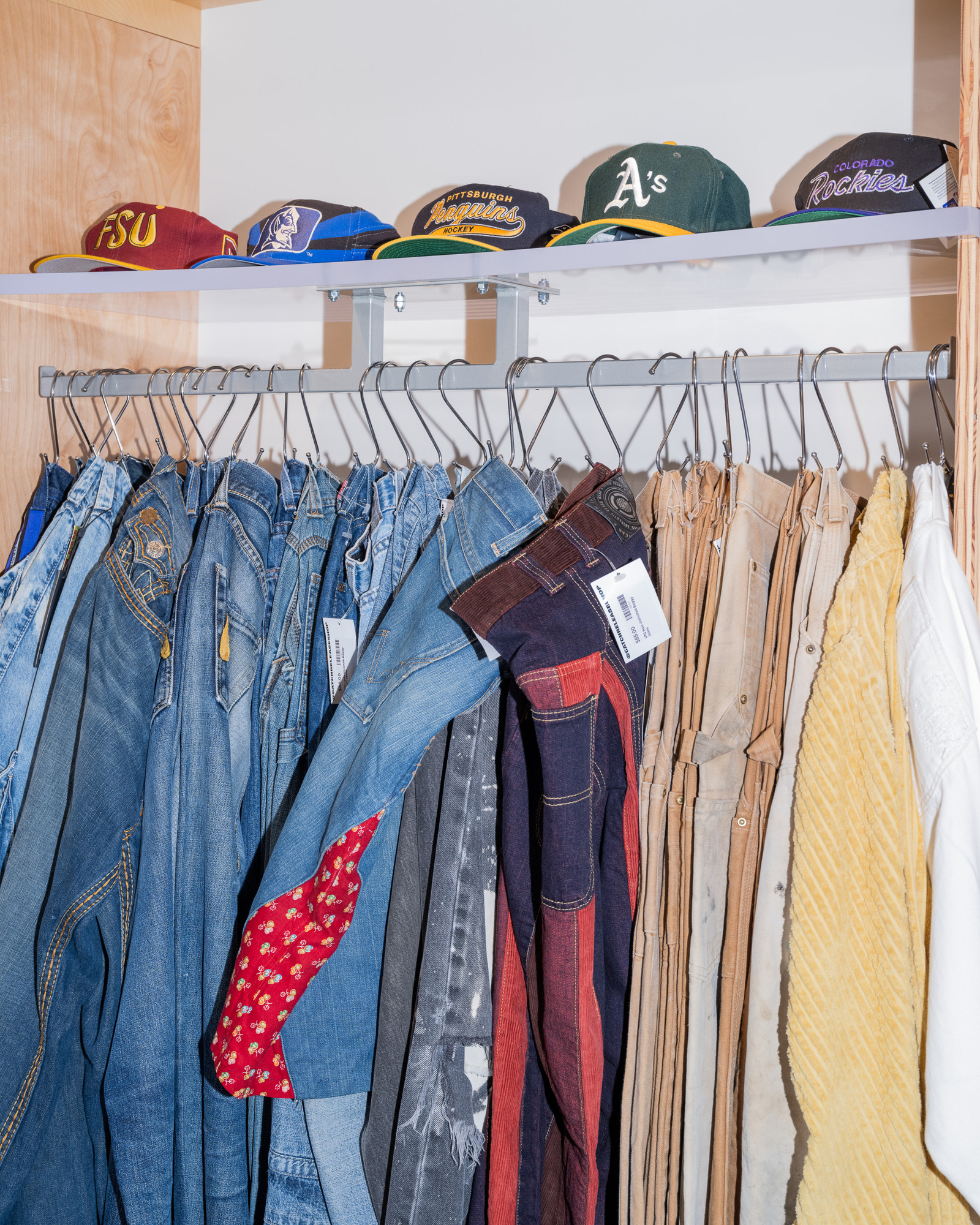 The image shows a row of colorful hats on a shelf above a rack of jeans and shirts hanging on hangers, with labels attached to the clothes.