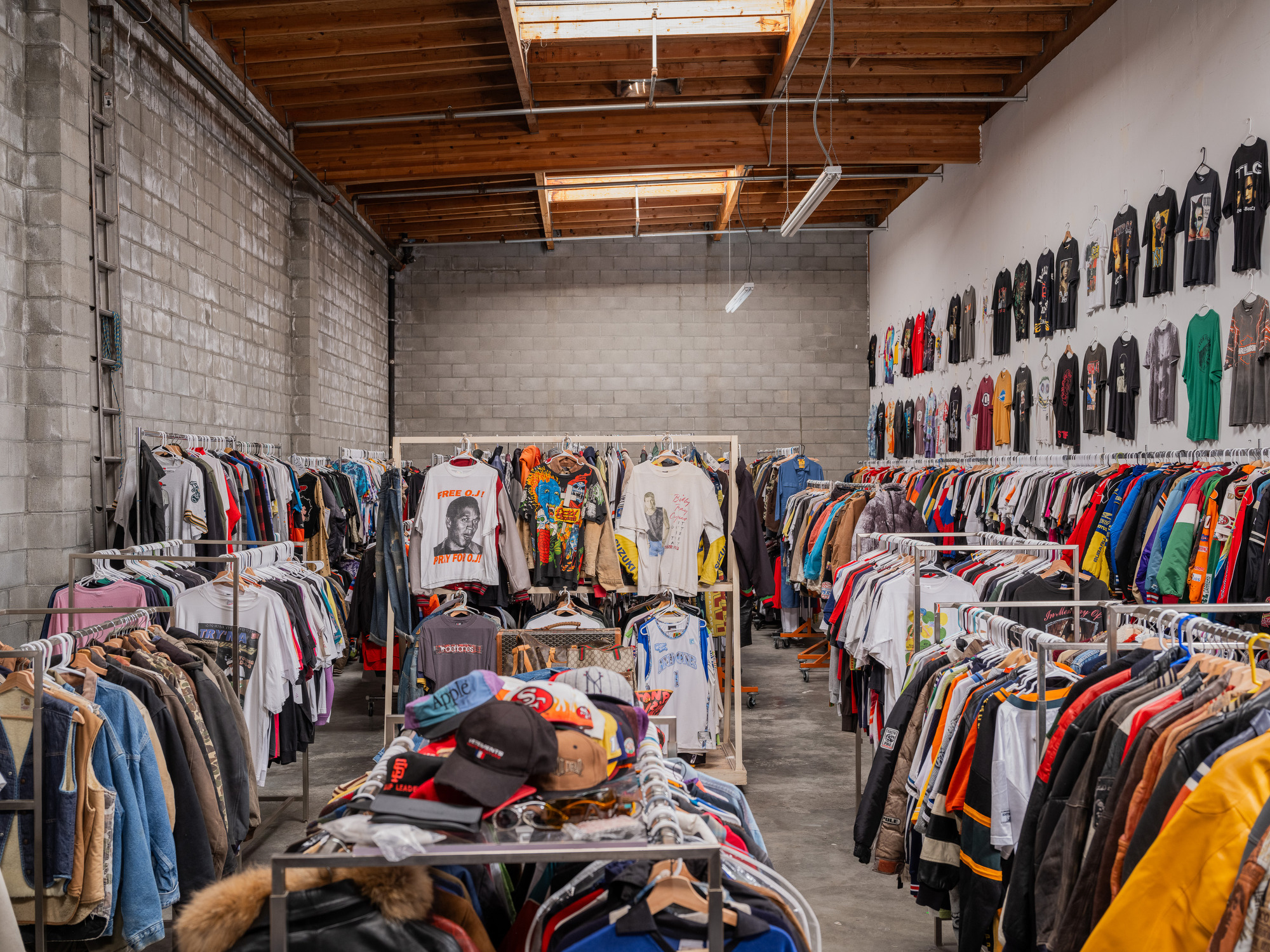 This image shows a vintage clothing store with rows of colorful shirts and jackets hanging on racks. The walls are lined with various T-shirts, and hats are piled on a table.