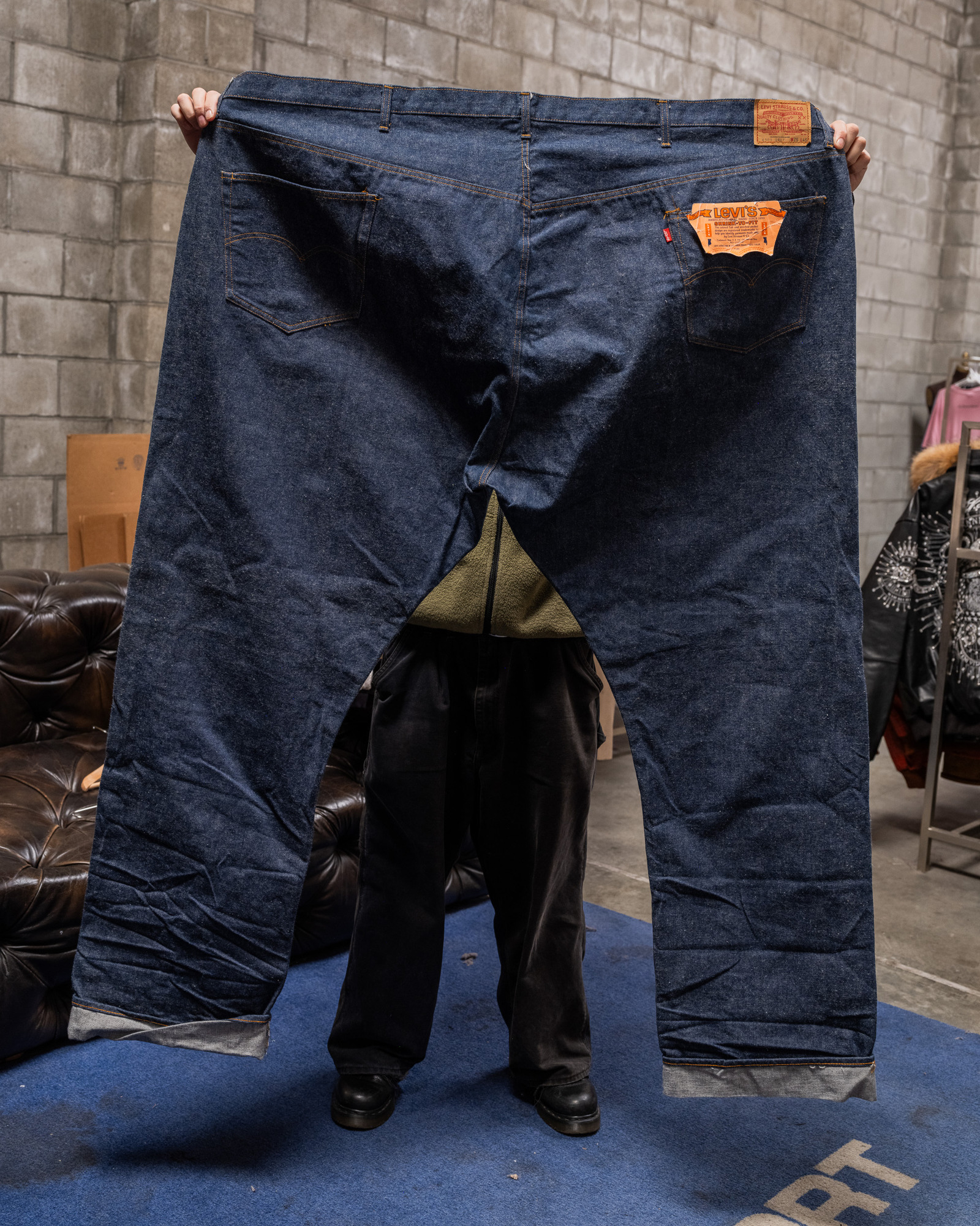 A person is holding up an enormous pair of blue jeans, wide enough to cover their entire body from view. The jeans appear comically oversized.