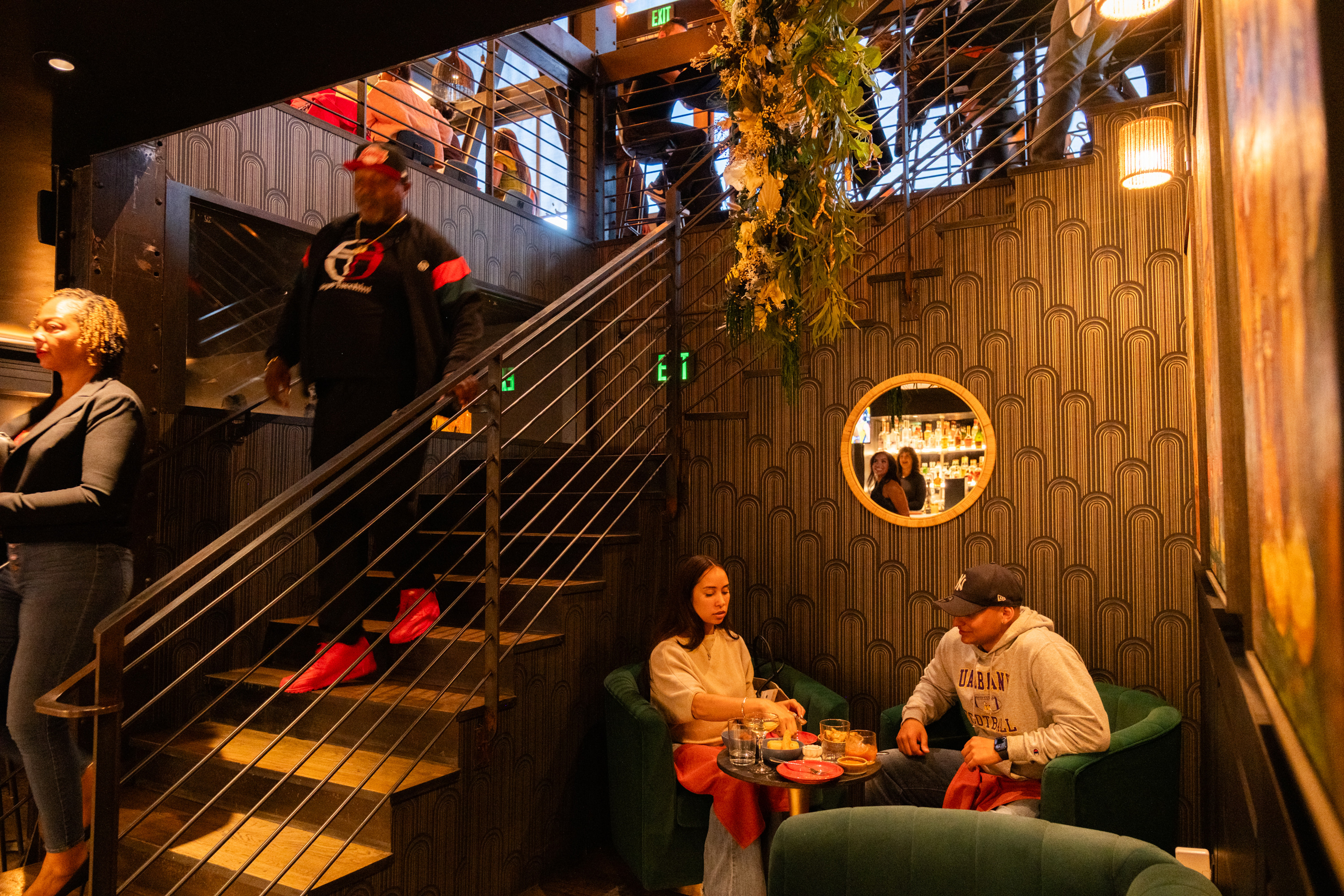 A stylish room features a staircase, patterned wallpaper, and a round mirror. Two people sit at a table with drinks, while others descend the stairs.