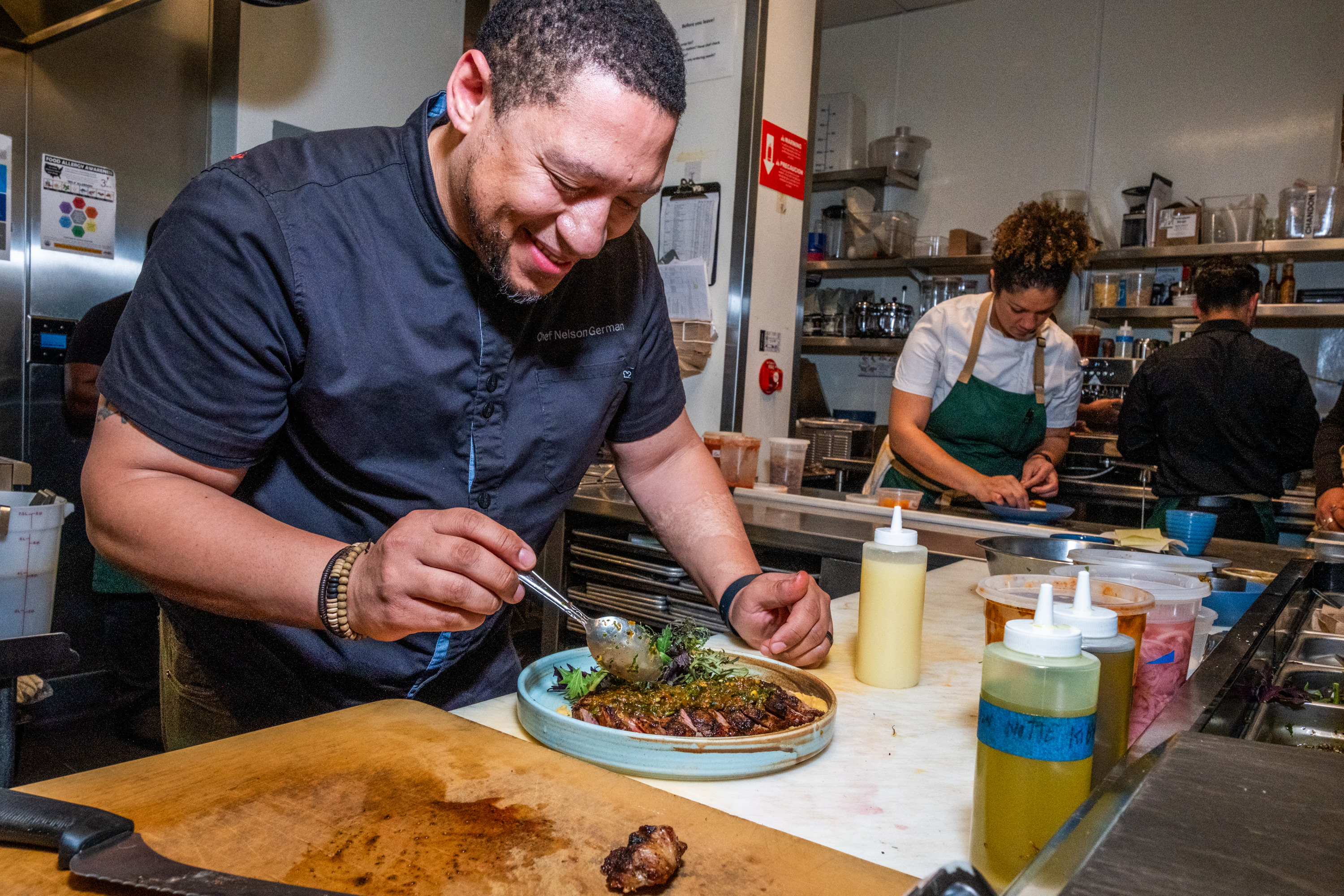 A chef smiles while carefully garnishing a plated dish in a busy restaurant kitchen. Other chefs work in the background, preparing ingredients.