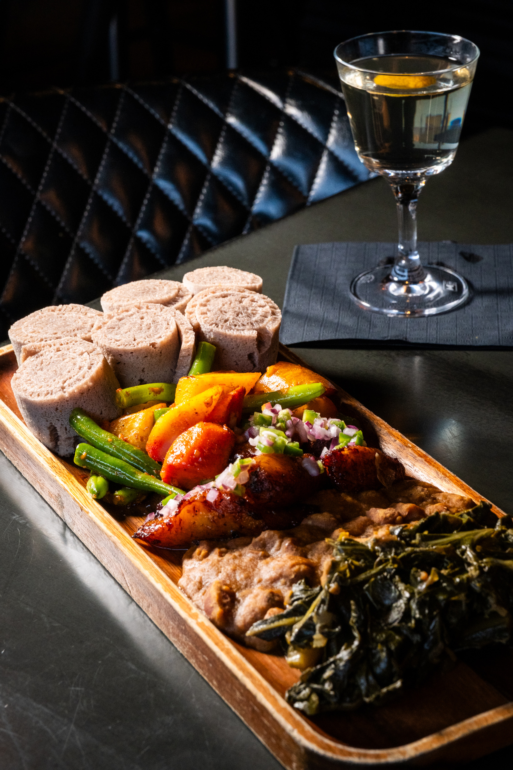 The image shows a wooden platter with rolled flatbread, assorted vegetables, cooked greens, and a meat dish, accompanied by a cocktail with a lemon twist.