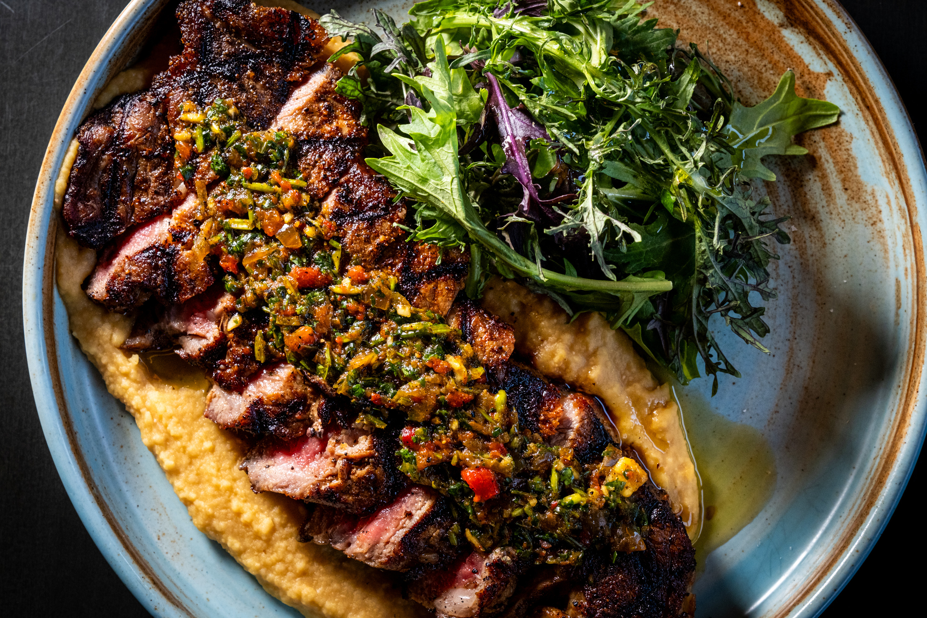 A charred sliced steak topped with colorful chimichurri sauce, resting on creamy mashed potatoes, alongside fresh mixed greens on a round ceramic plate.