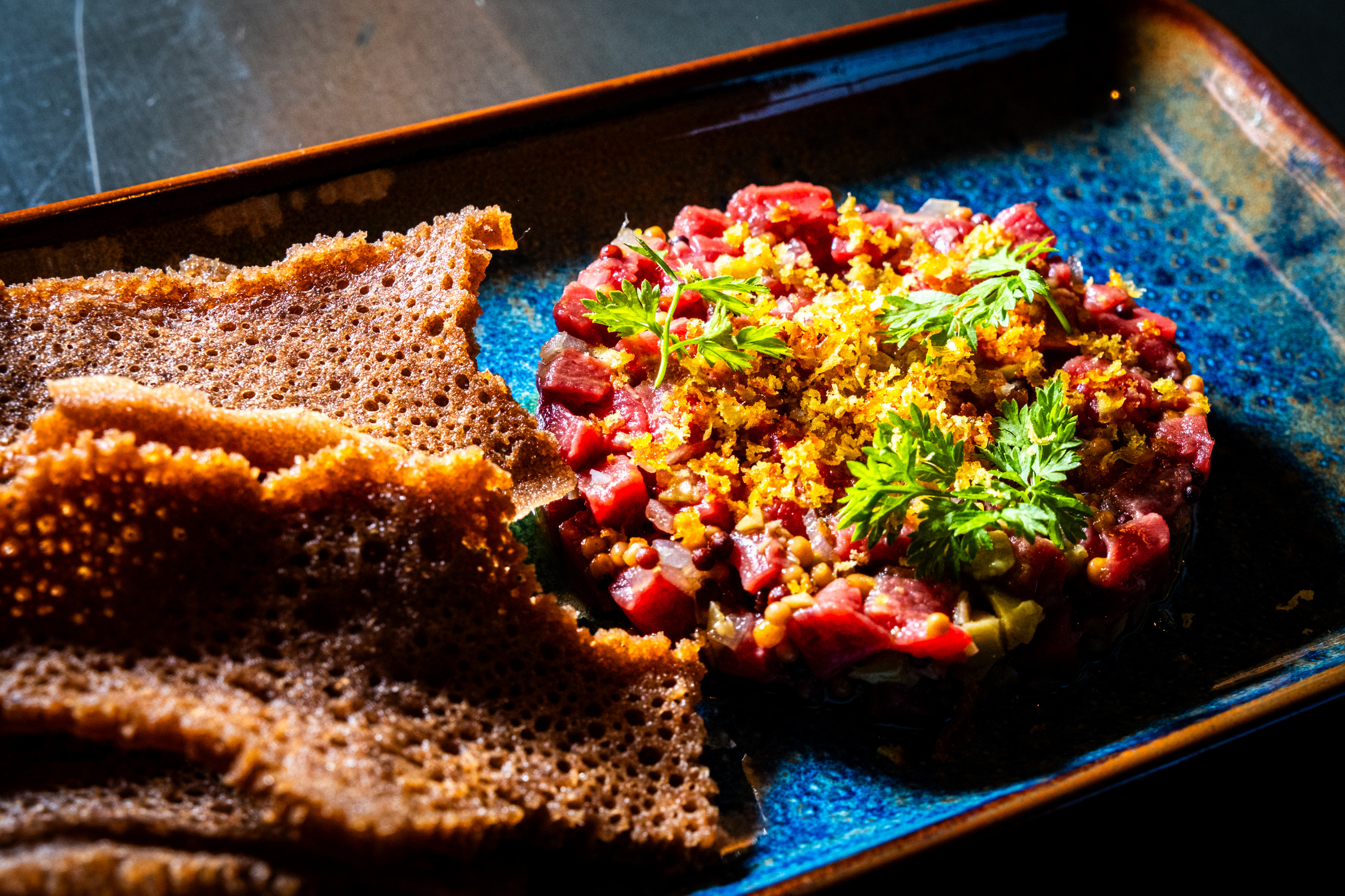 A vibrant dish with diced red meat topped with herbs and crispy bits, accompanied by crispy, brown-edged flatbreads on a blue rectangular plate.