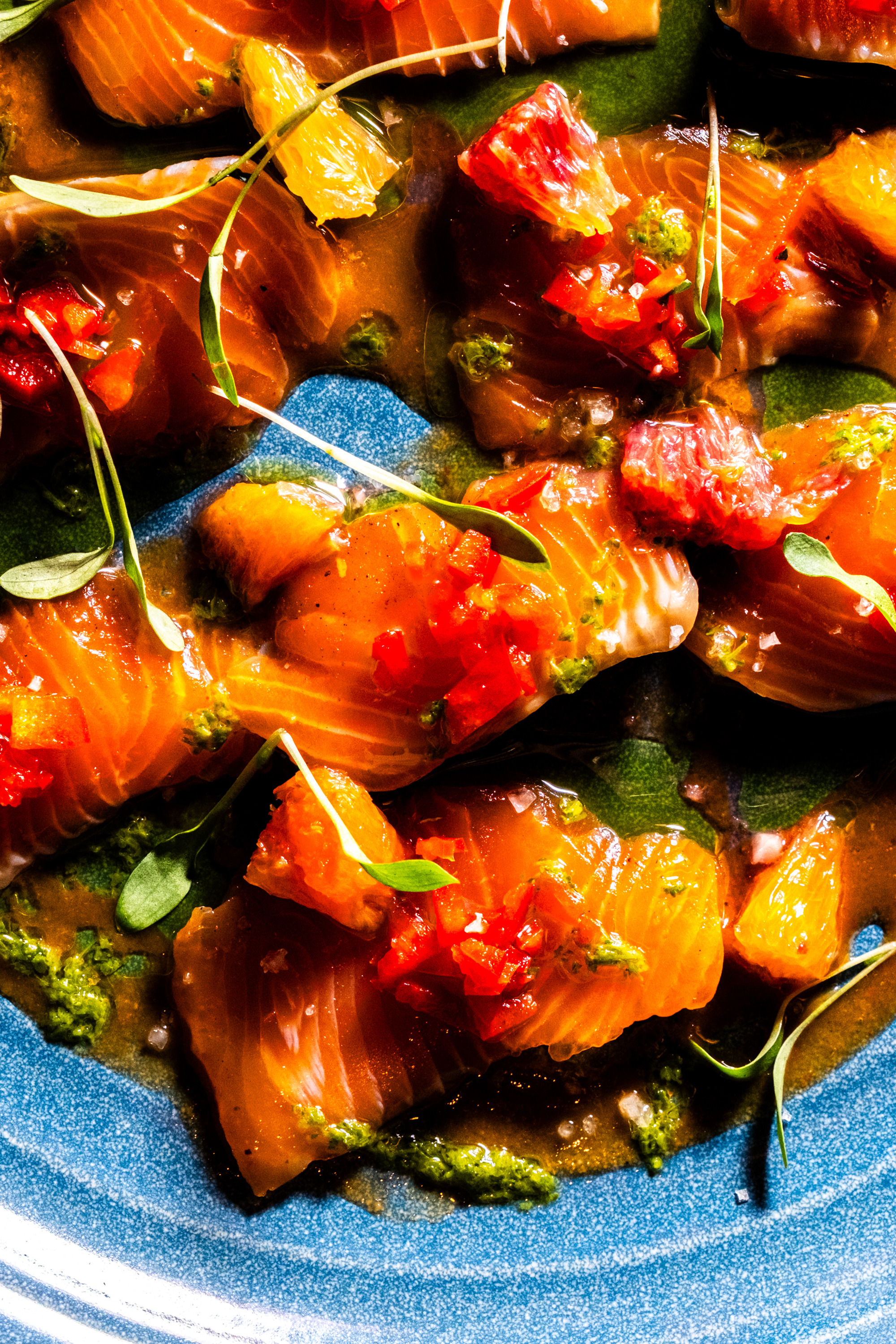 Slices of raw fish are artfully arranged on a vibrant plate, garnished with small greens, diced red peppers, and citrus segments, all drizzled with a green sauce.