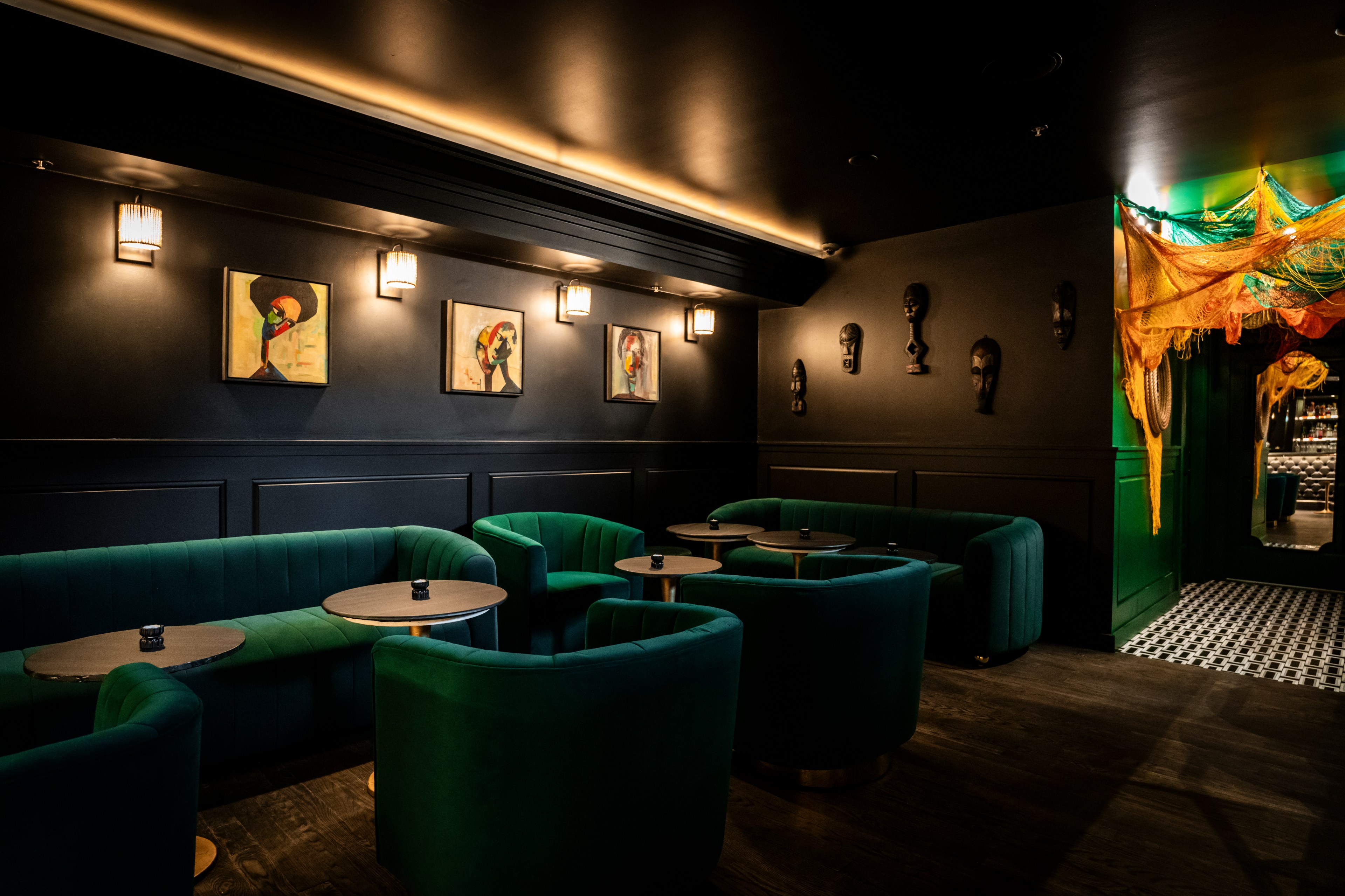 The image shows a dimly lit lounge with green velvet seating, small round tables, and modern artwork on dark walls. Masks and colorful netting adorn the space.