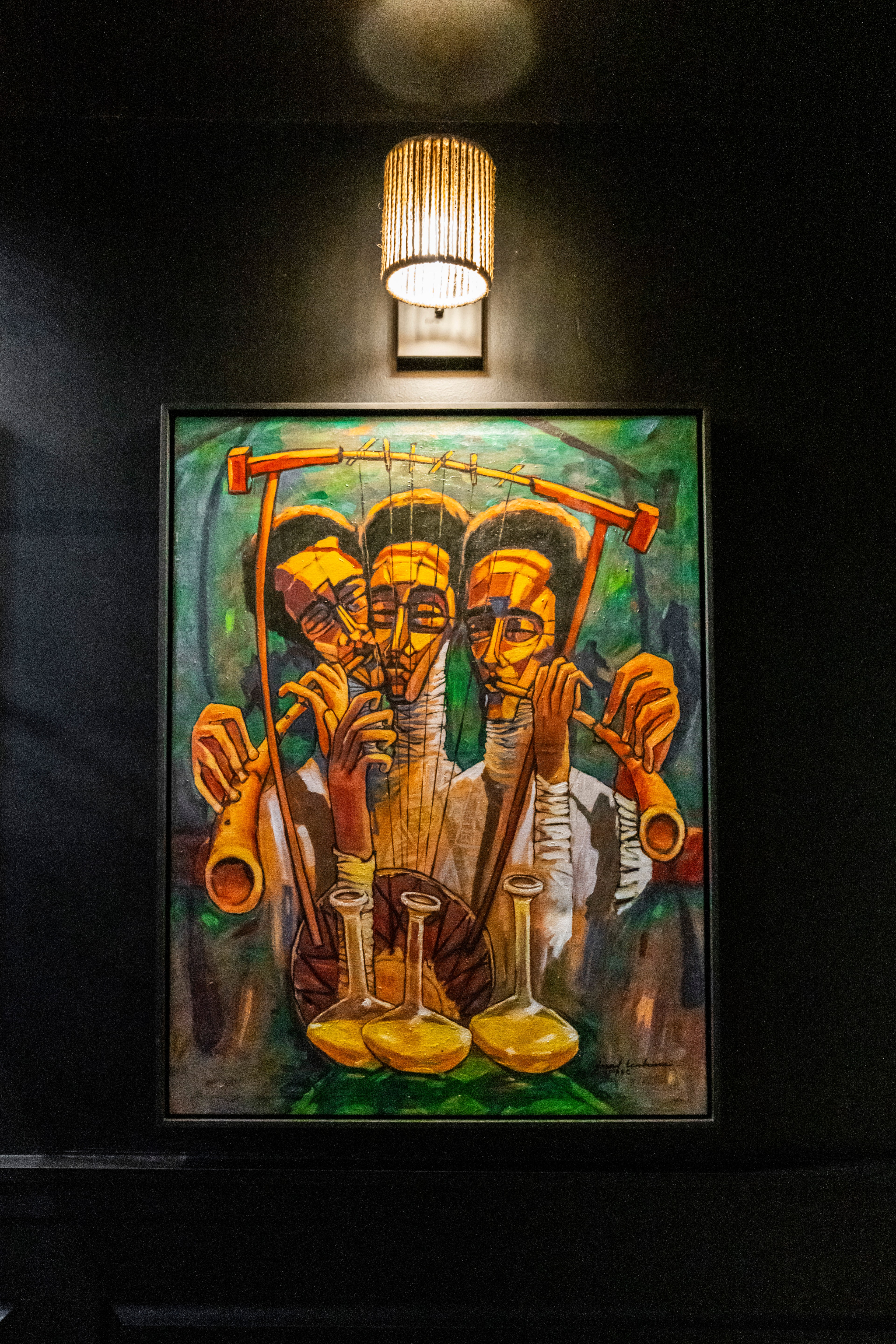 The image shows a colorful painting of three abstract figures playing musical instruments, under a warm, lit sconce on a dark wall background.