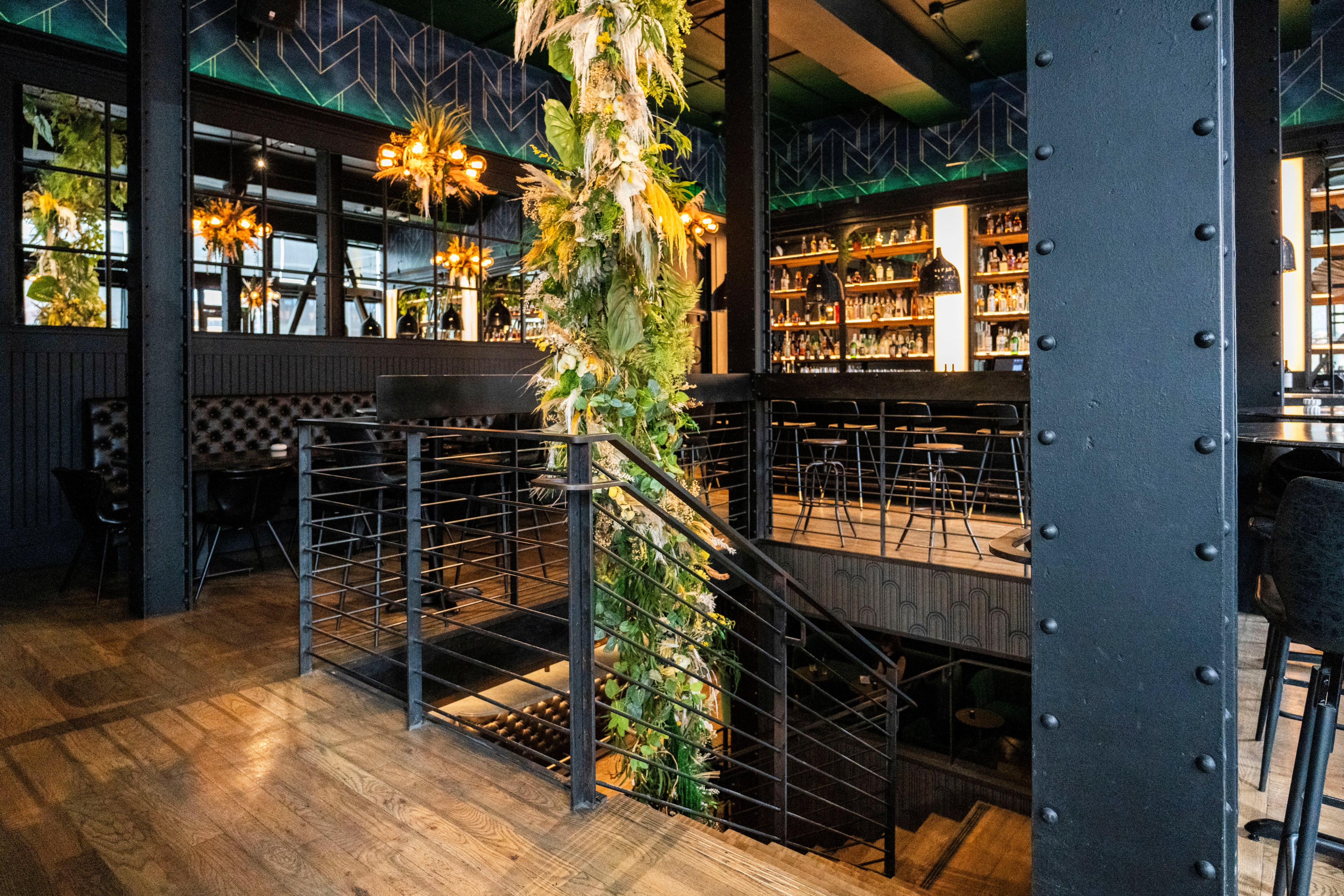 The image shows a stylish, modern bar with a staircase, decorated pillars with greenery, warm lighting, and a stocked bar with high stools.