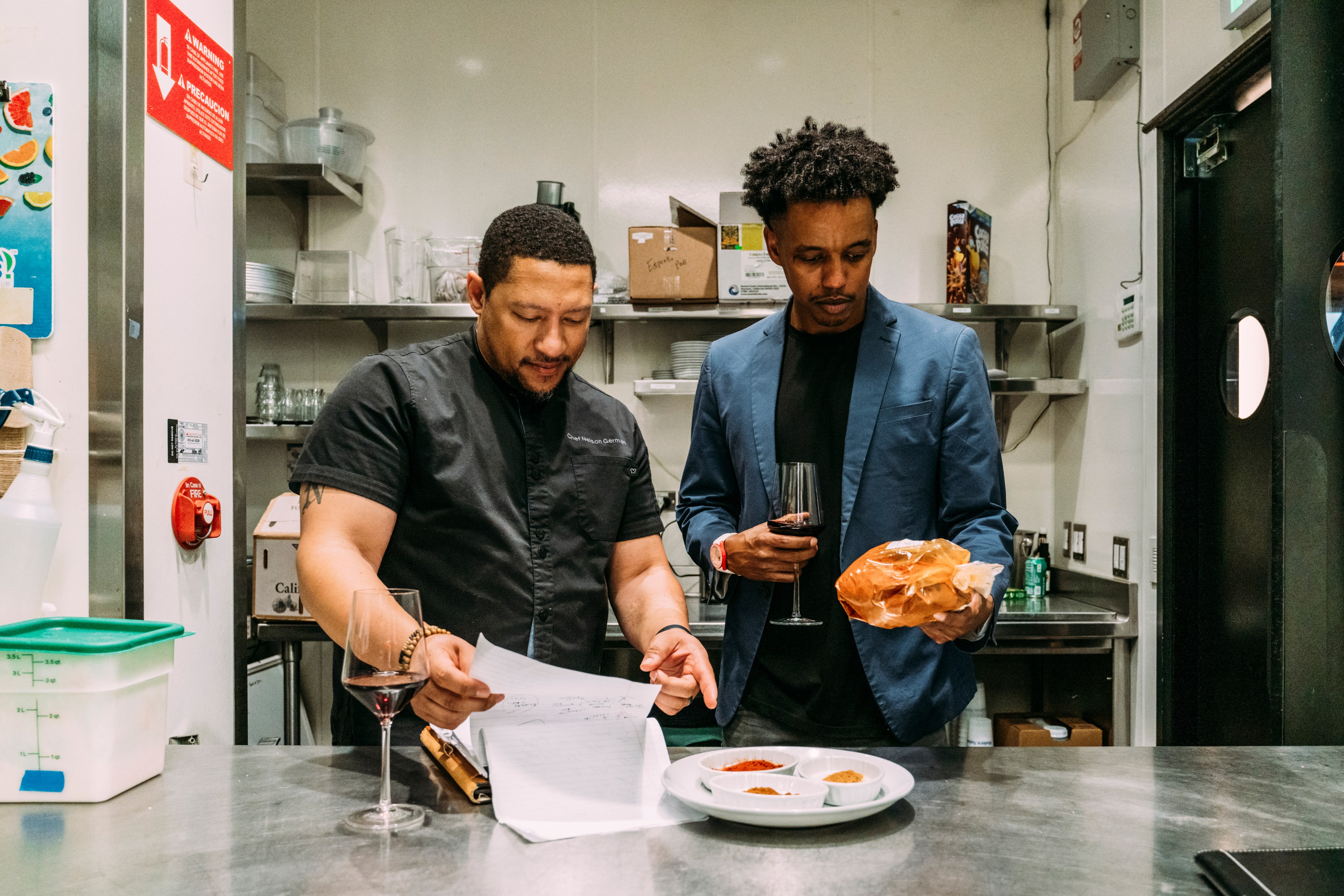 Two men stand in a kitchen, one holding papers and the other holding bread and a wine glass. Spices in small bowls are on the counter.