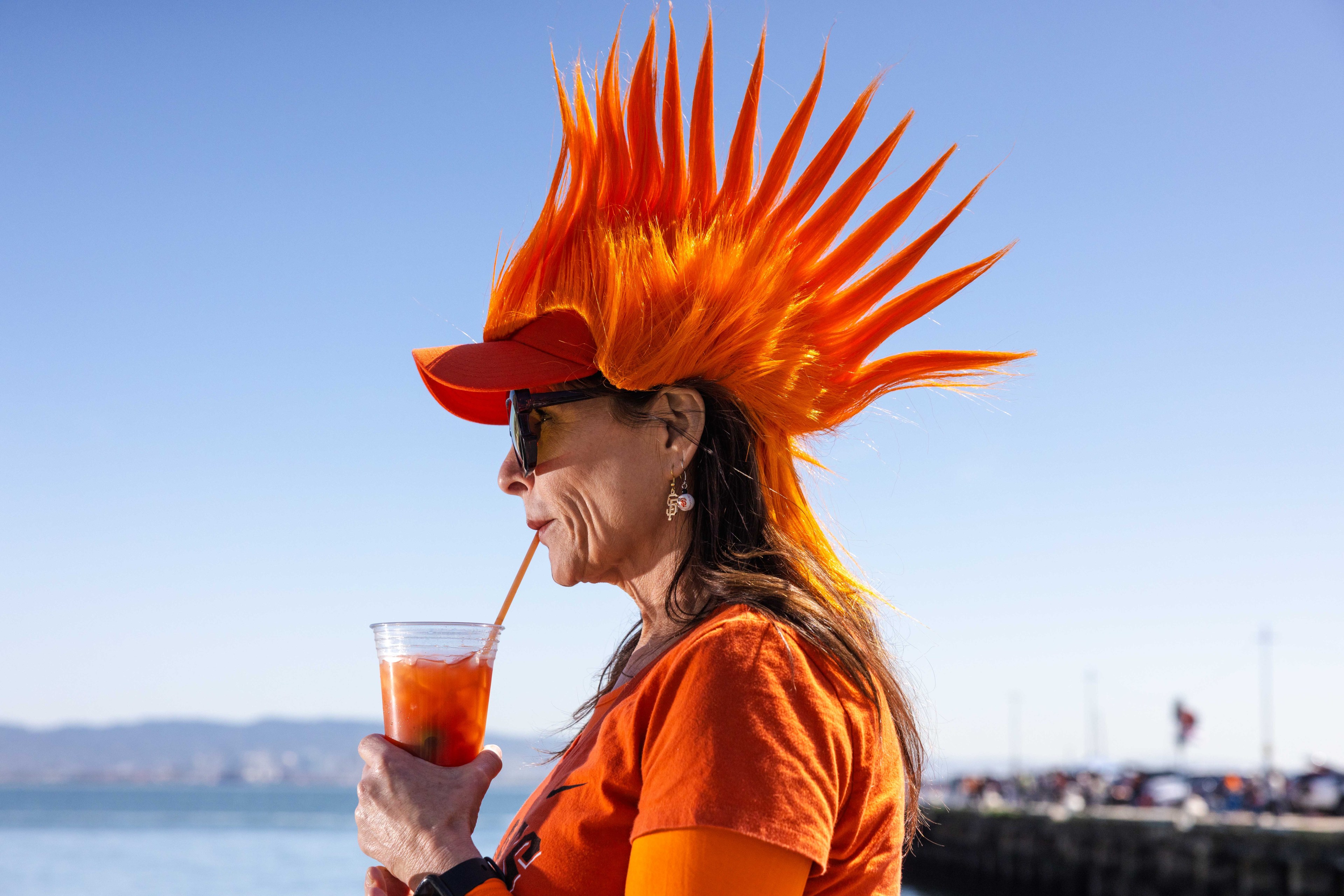 The image shows a person wearing a bright orange spiky wig and matching outfit, sipping a drink through a straw. They're outdoors, with a clear blue sky.