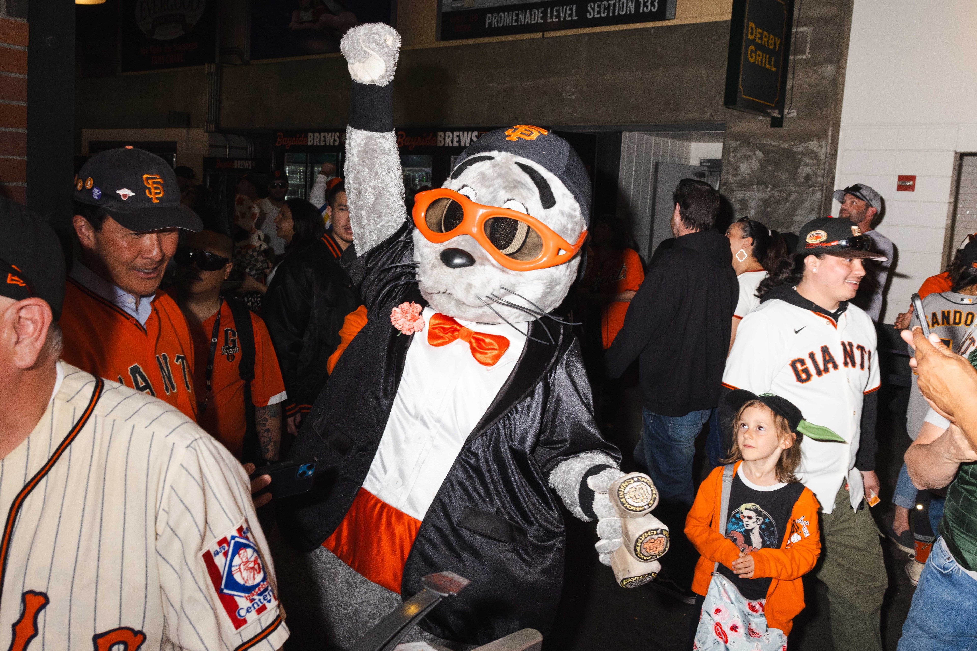 A mascot in a tuxedo and orange sunglasses raises a fist among people in San Francisco Giants attire. The crowd seems excited and engaged.
