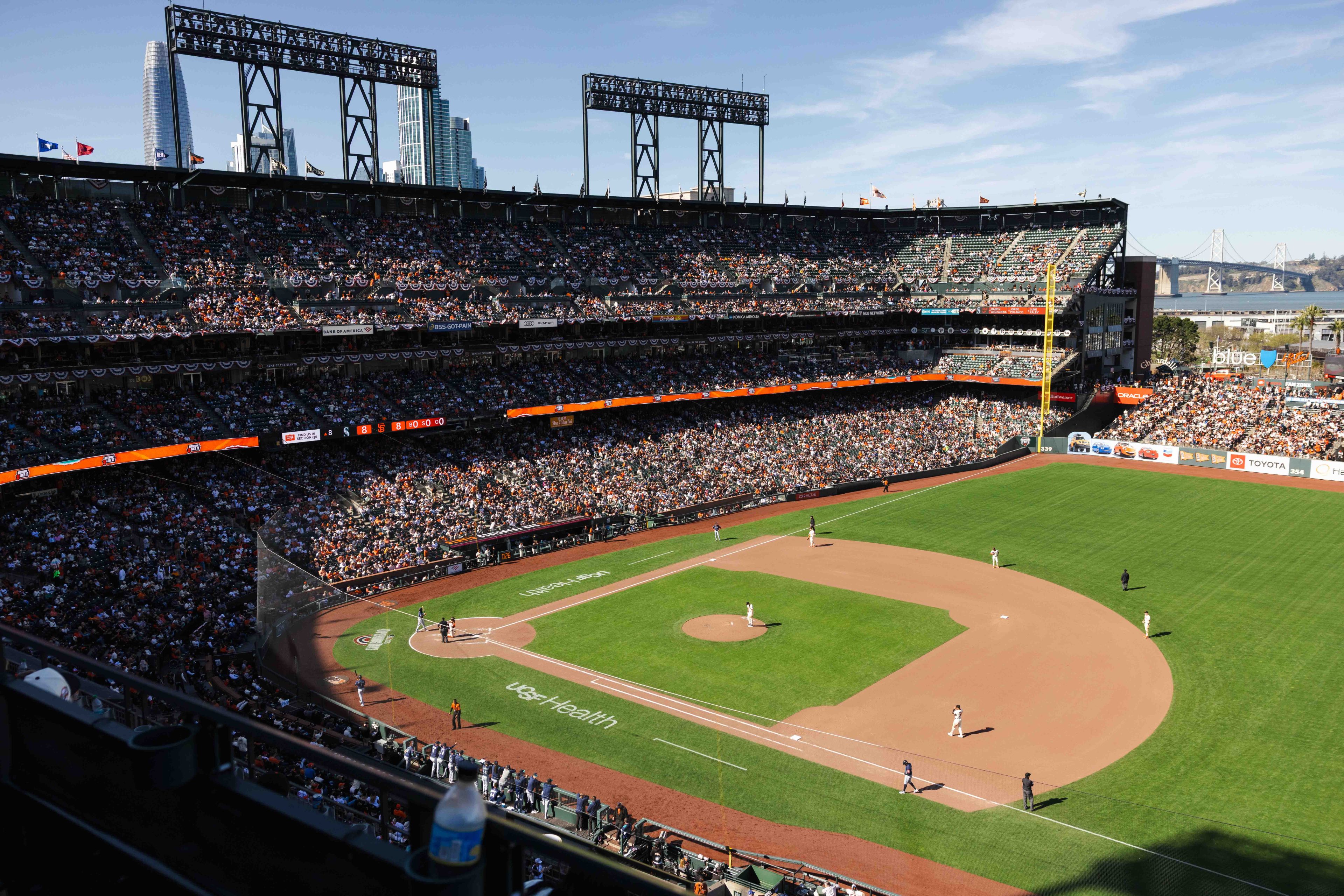 A packed baseball stadium with players on the field during a sunny day. The field is lush and green, with cityscape and a bridge visible in the background.