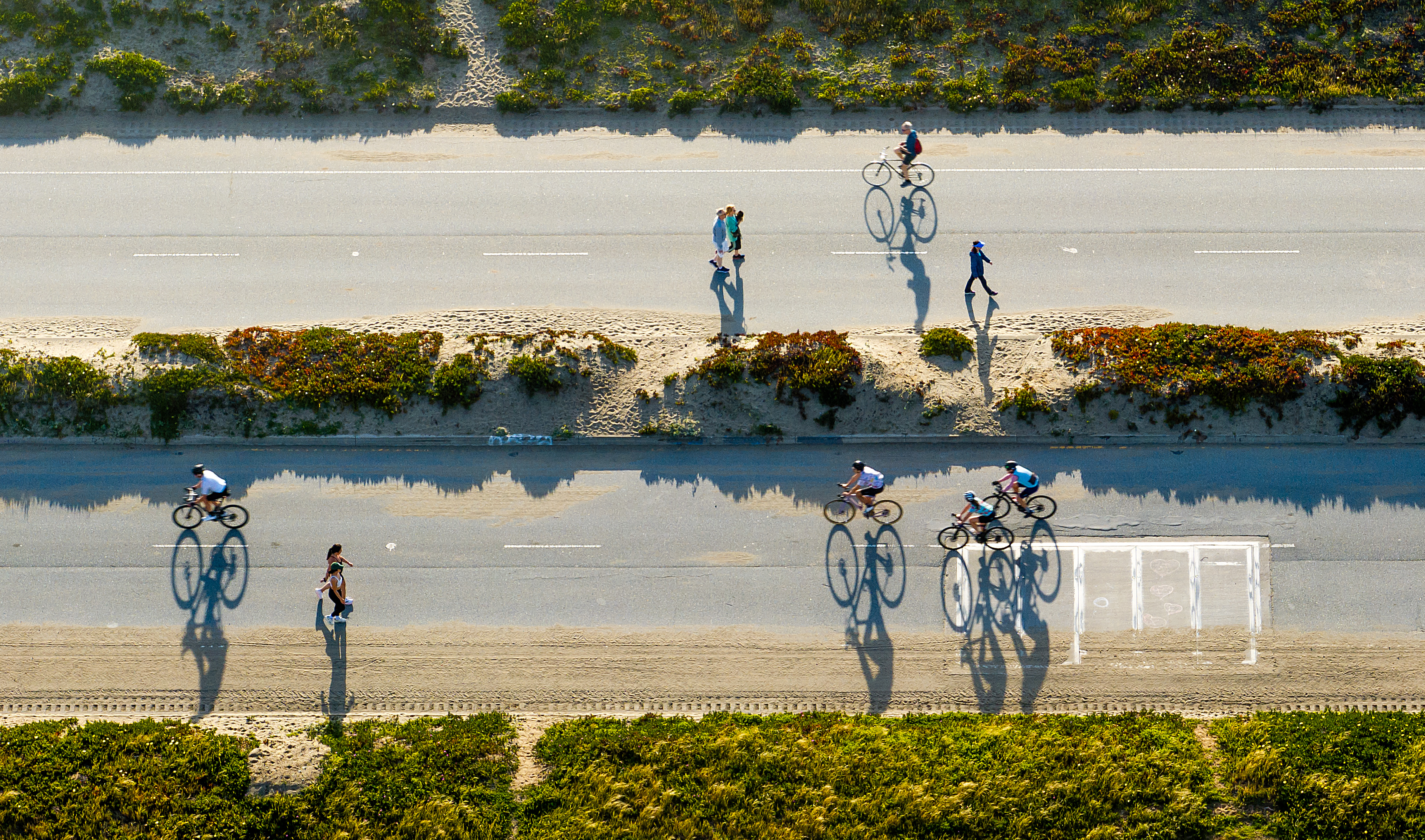 The image shows an aerial view of a road with cyclists and people walking, casting long shadows on the pavement. There's vegetation lining the road.