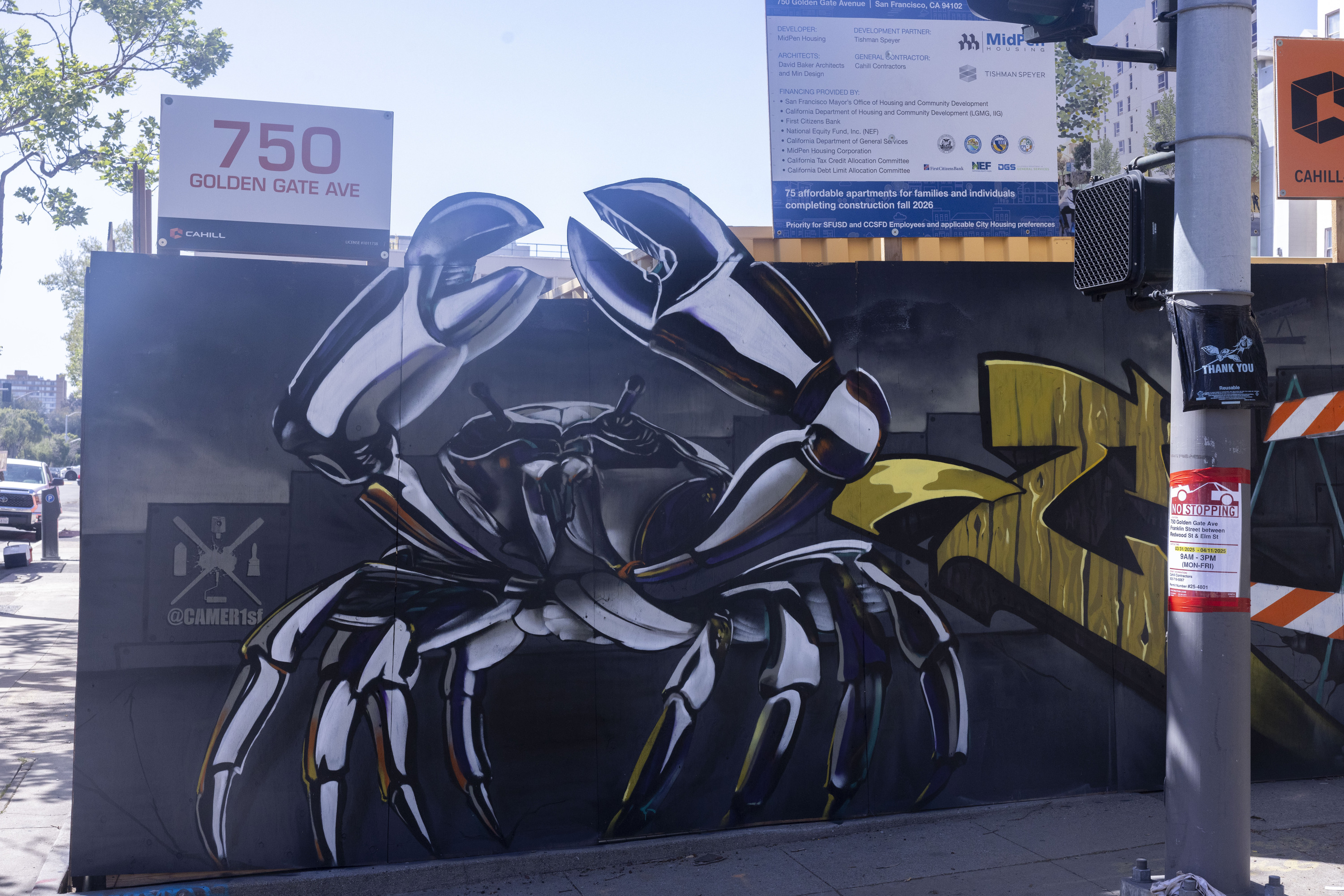 A large street mural depicts a detailed, futuristic crab with metallic claws. It’s painted on a temporary construction wall with various signage above.