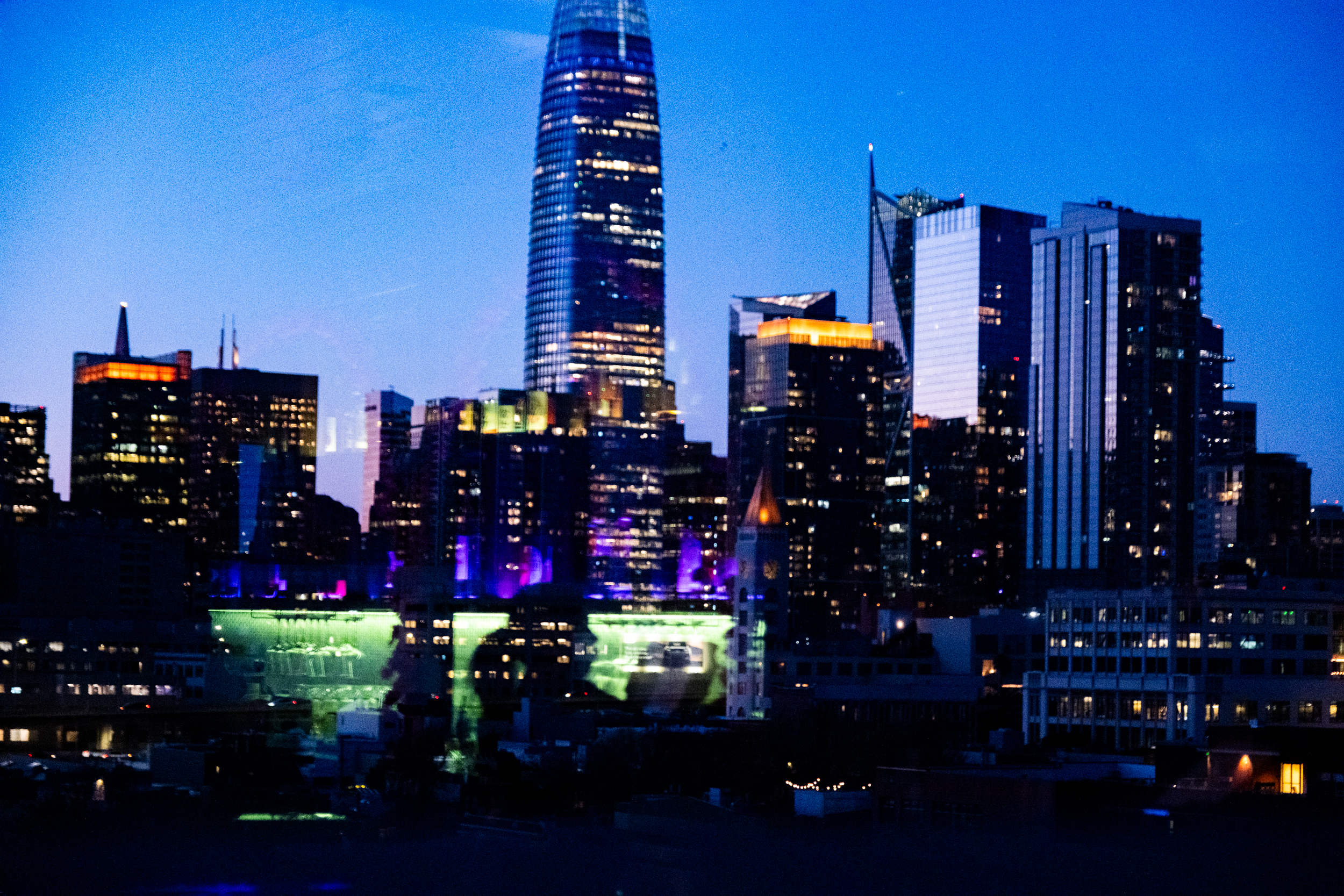 The image shows a vibrant city skyline at dusk with tall buildings illuminated by colorful lights against a deep blue sky.
