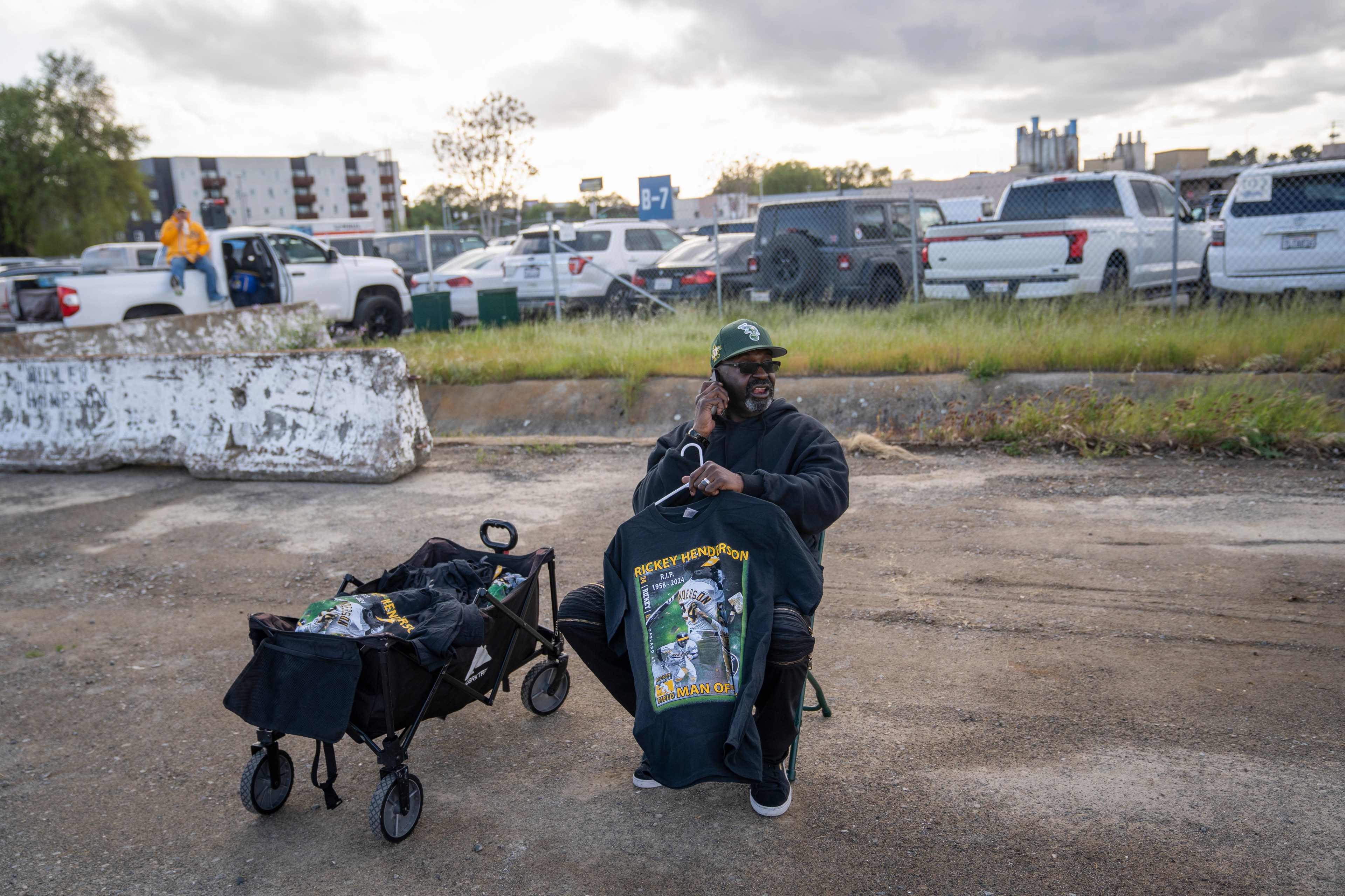 A man sits on a chair in a parking lot, holding up a black T-shirt with a design. A cart beside him holds more shirts, and there are cars parked in the background.
