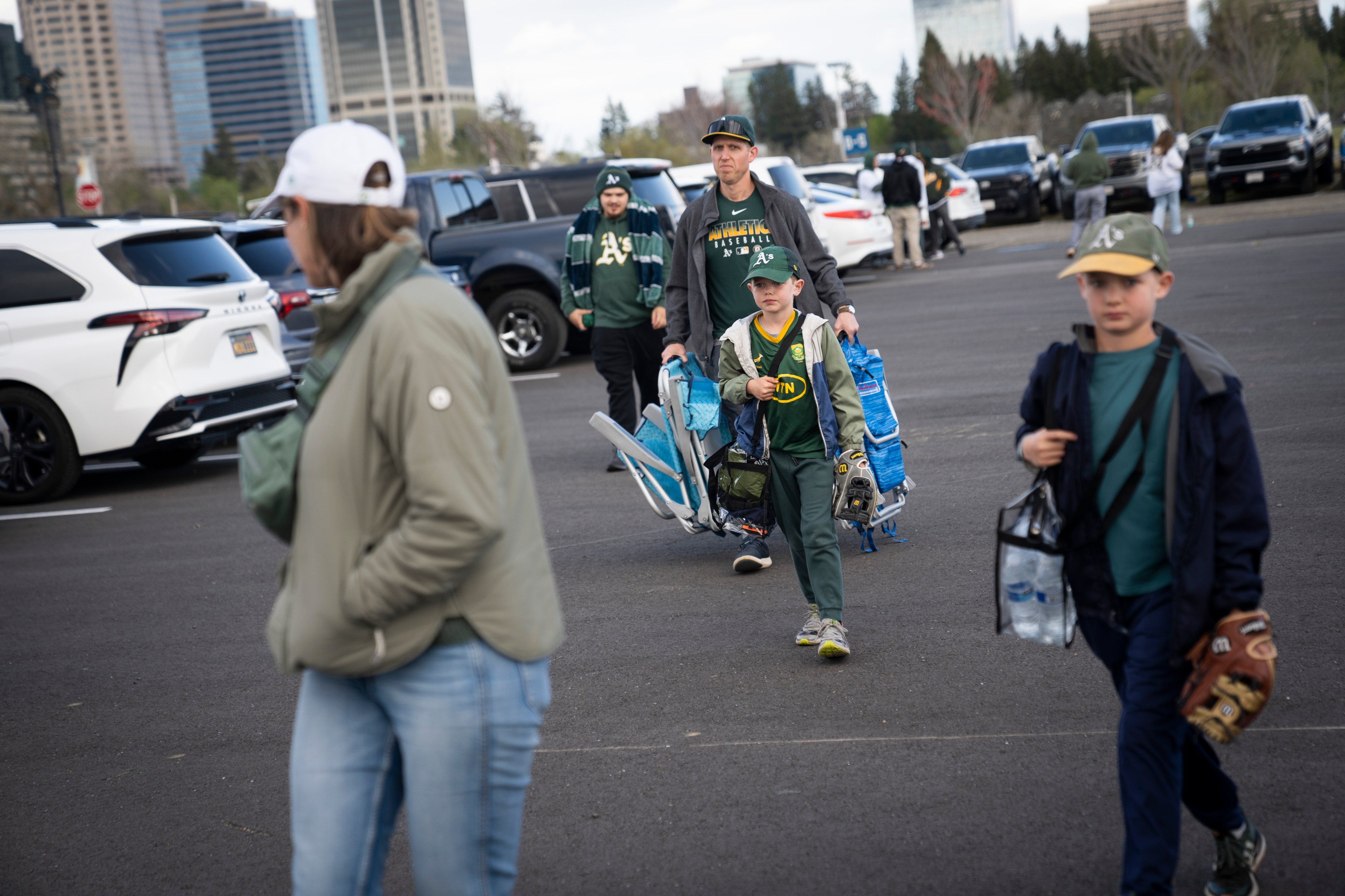 Several people walk through a parking lot carrying baseball gear. They are wearing green and yellow apparel. Cars and a city skyline are in the background.