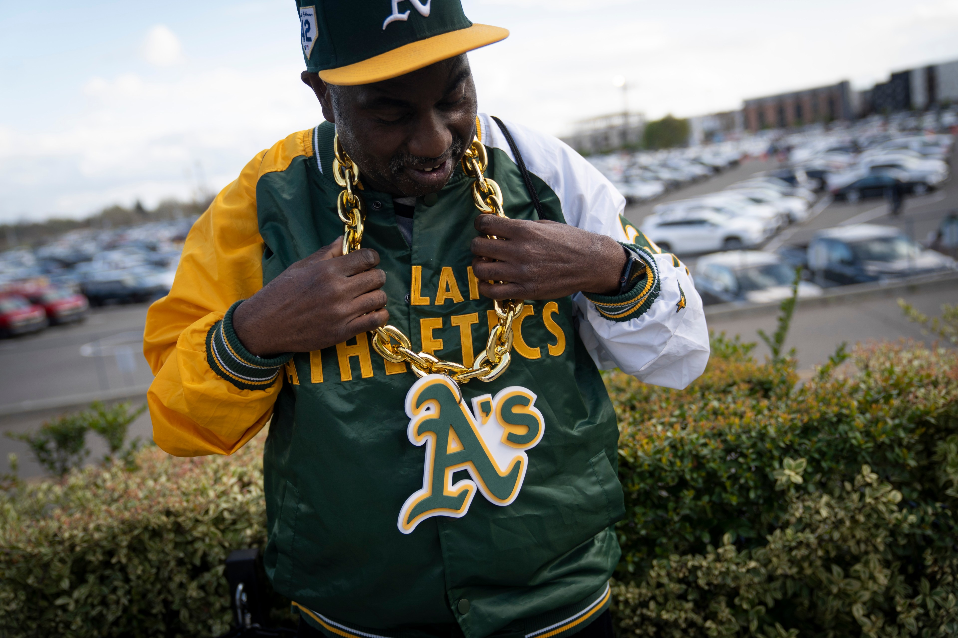 A man in an Oakland Athletics jacket and cap wears a large gold chain with the team's logo. He's outdoors with a parking lot in the background.