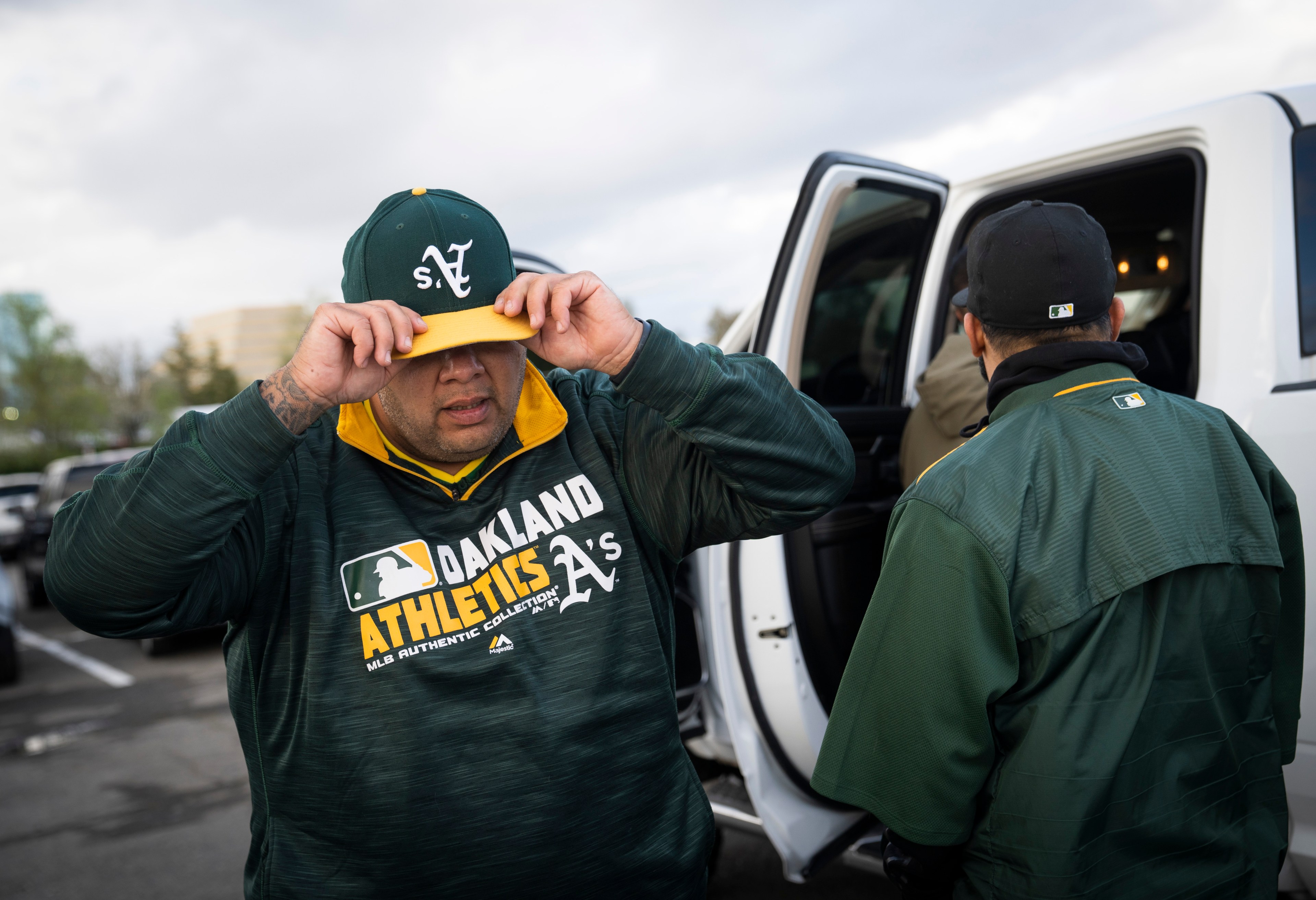 A person adjusts a cap labeled &quot;SV&quot; and wears an Oakland Athletics sweatshirt, standing next to a white vehicle with another person nearby.