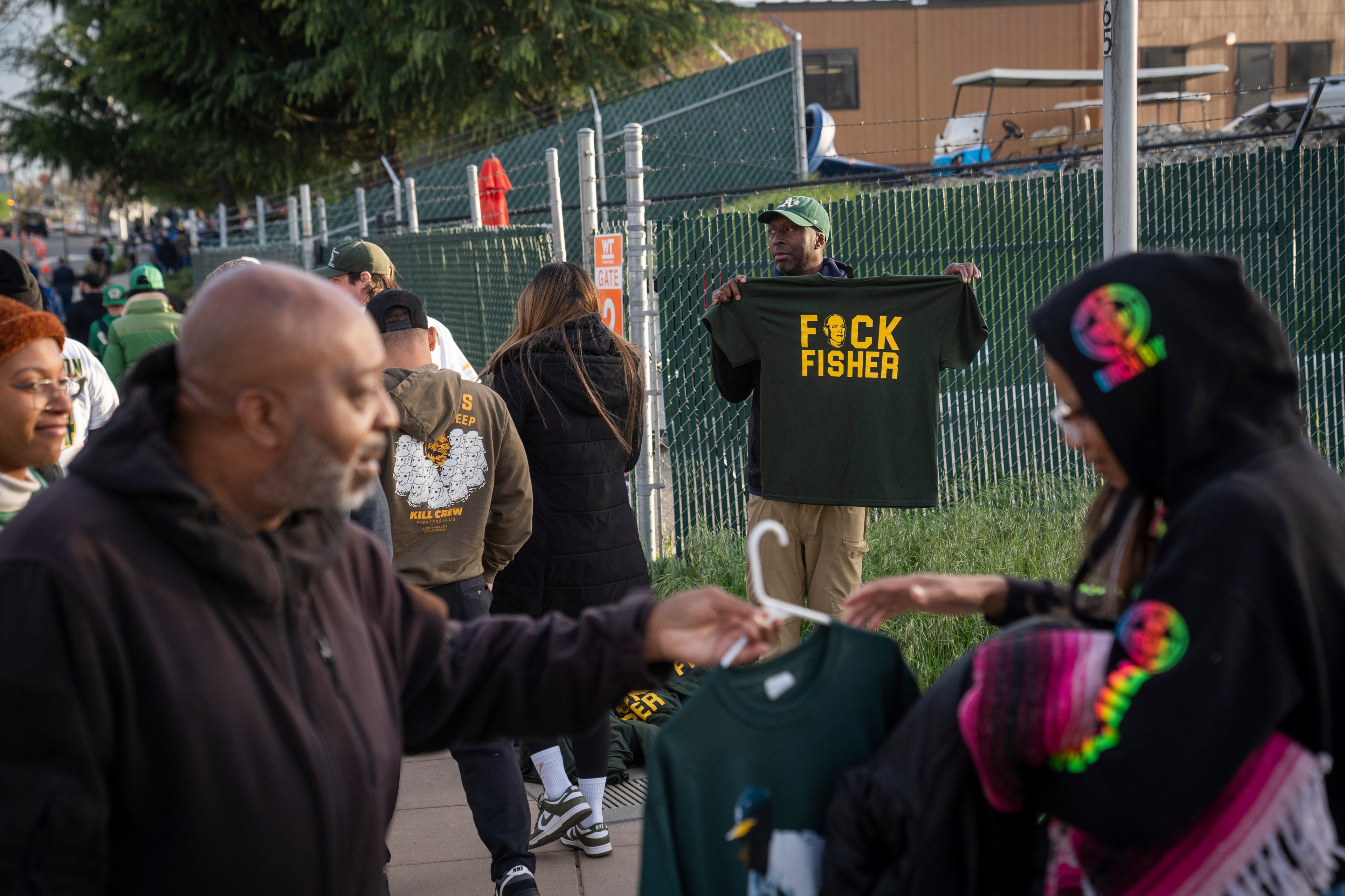 People stand near a fence at an outdoor market, including a man holding a green shirt with yellow lettering. Others browse nearby merchandise, with trees in the background.