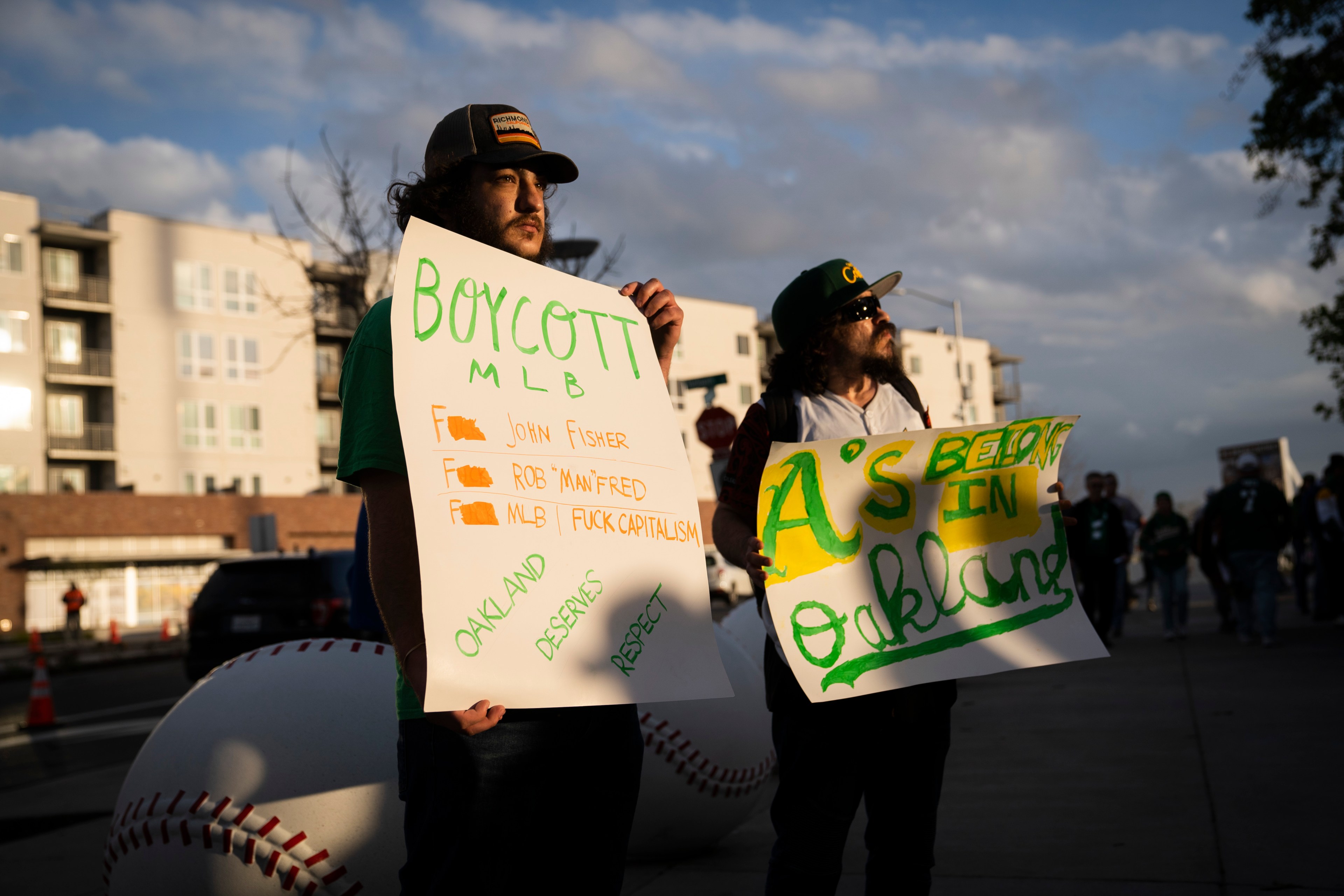 Two people hold protest signs. One reads &quot;Boycott MLB,&quot; listing grievances, and the other says &quot;A's Belong in Oakland.&quot; They're standing on a street.
