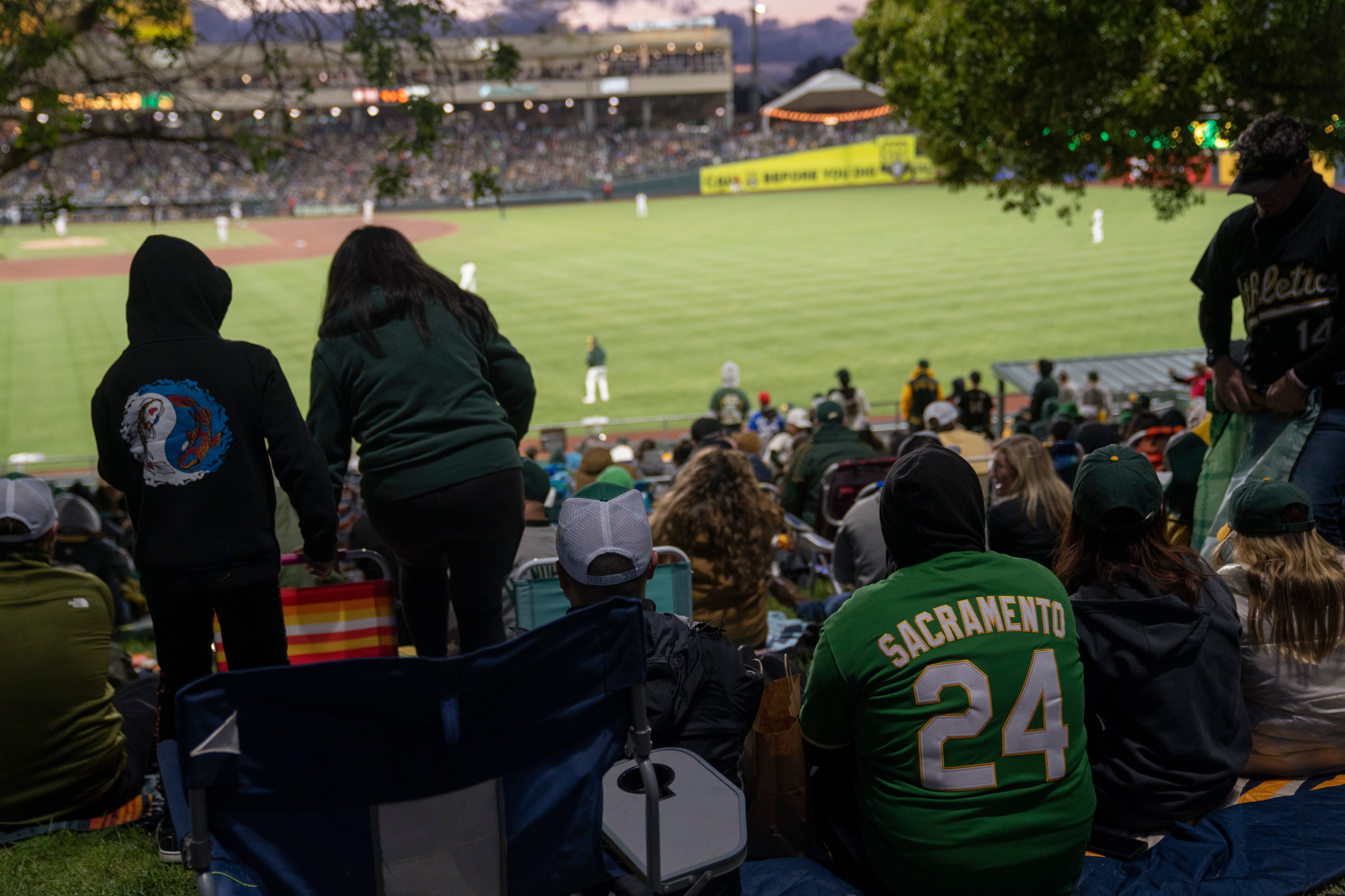 A crowd of people on a grassy hill watches a baseball game. Many wear green, including one with &quot;Sacramento 24&quot; on their jersey. The stadium is well-lit in the background.