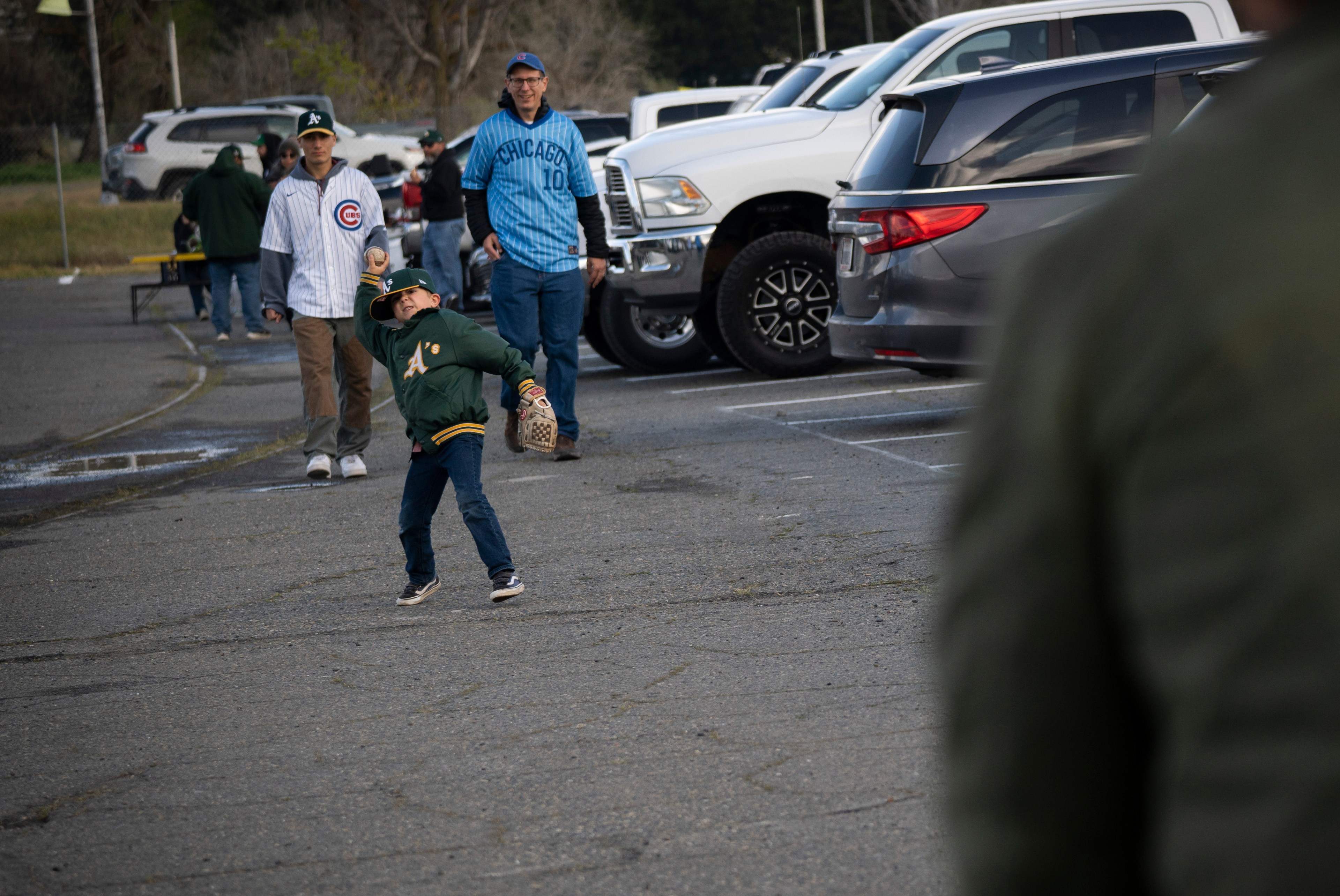 A child in an A's jacket throws a baseball in a parking lot, while two adults in sports jerseys watch near parked cars.