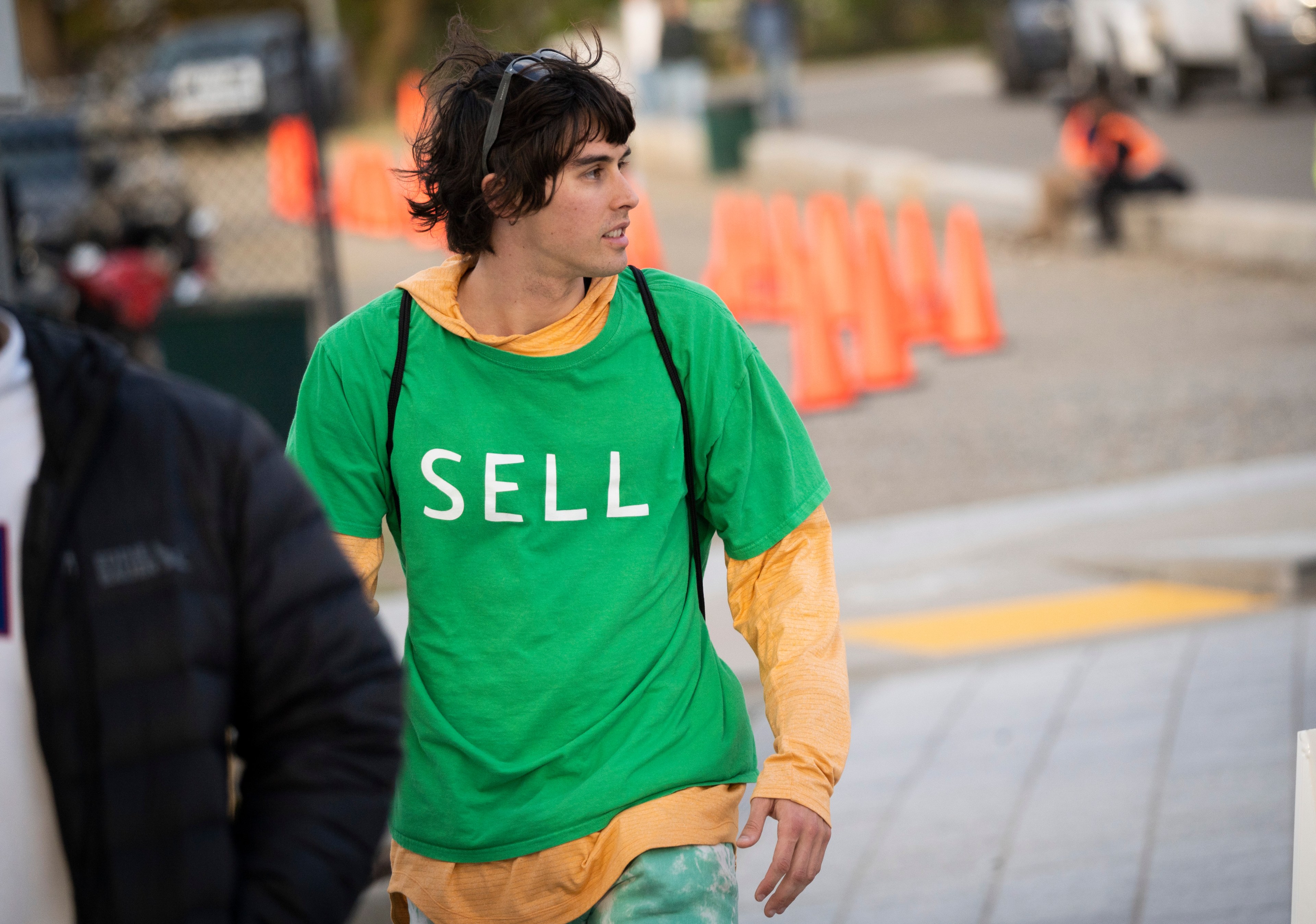 A person in a green shirt with &quot;SELL&quot; printed on it is walking outdoors. There are orange traffic cones and blurred figures in the background.