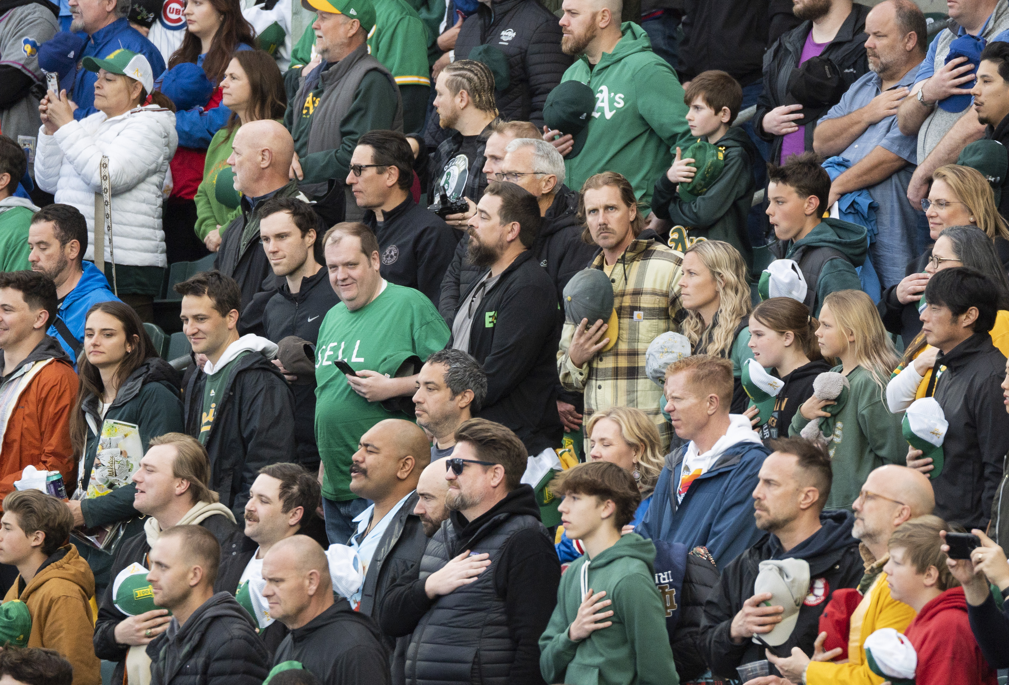 The image shows a crowd of people at a sports event, standing with hands over their hearts. They wear various team apparel, indicating a shared enthusiasm.