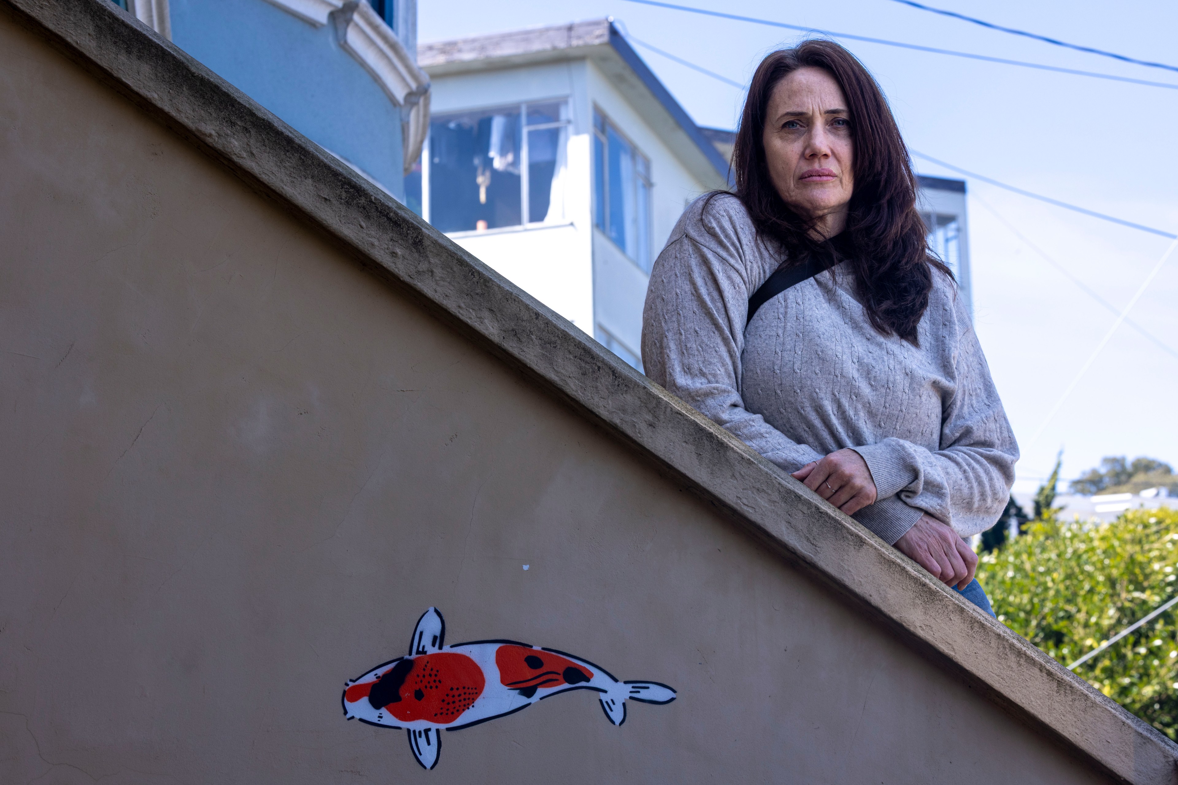 A woman leans on a sloped wall with a koi fish mural. She wears a gray sweater, and the background shows blue sky and buildings.