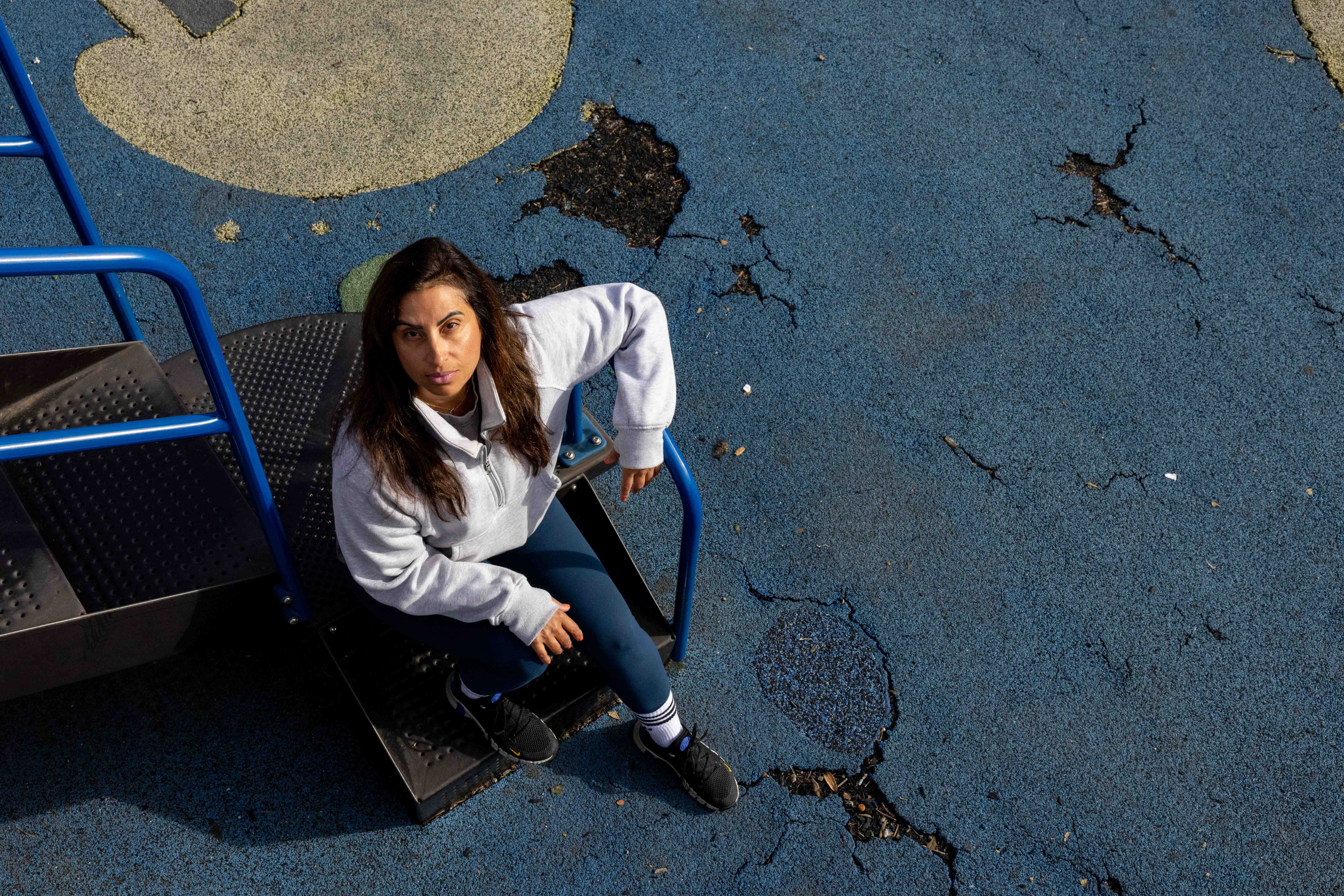 A woman sits on playground steps, looking up towards the camera. She's wearing a light sweatshirt and dark leggings. The ground around her is blue and cracked.
