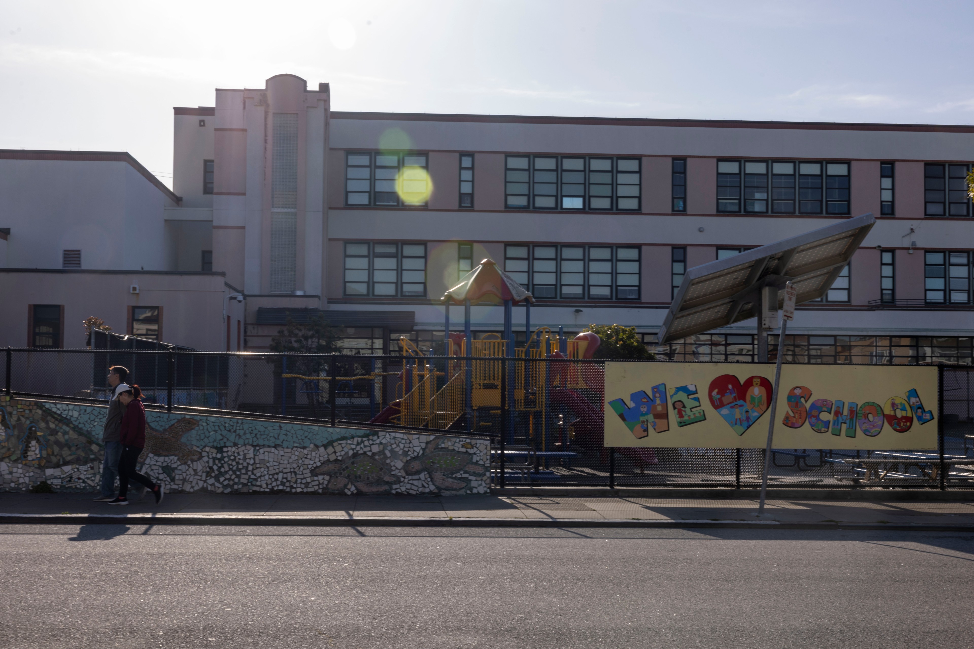 A school building with large windows is in the background. A colorful playground is behind a fence with a &quot;We Love School&quot; banner. Two people walk past.