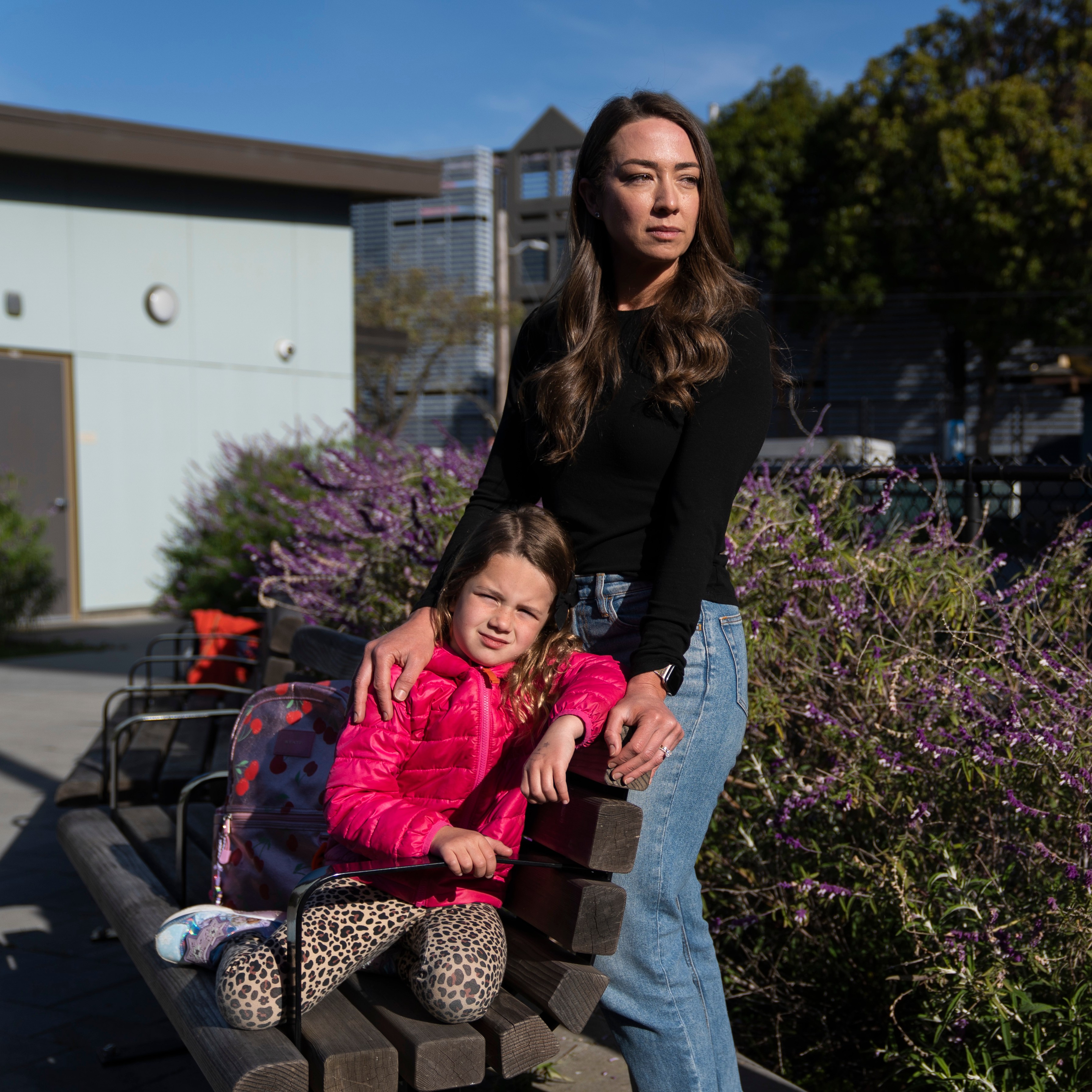 A woman in a black top and jeans stands next to a sitting child in a pink jacket and leopard-print leggings. They're outdoors near purple flowers.