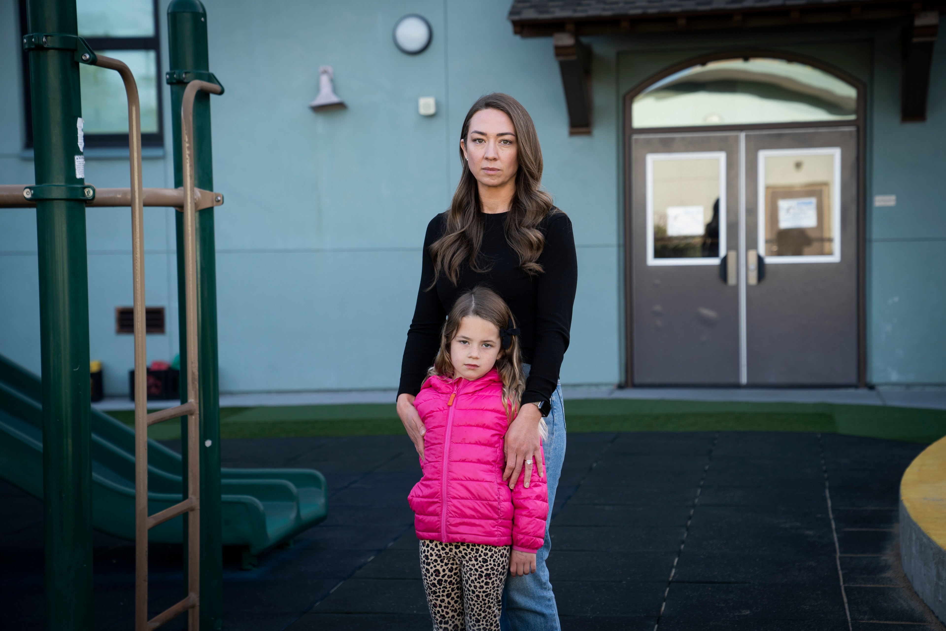 A woman in a black top stands next to a young girl in a bright pink jacket and leopard print leggings. They're in front of a playground and a building entrance.