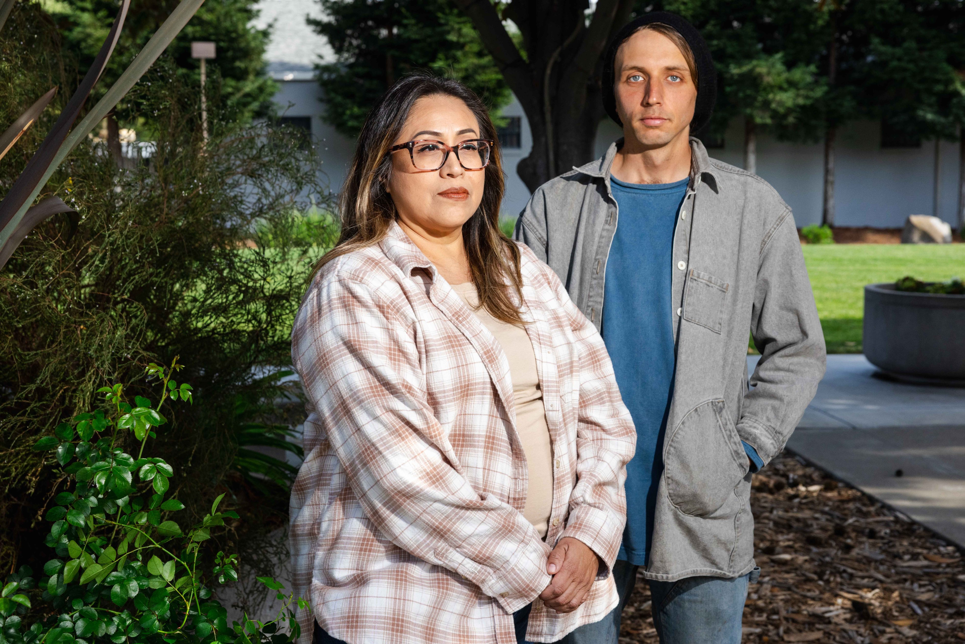 A woman and a man stand outdoors, both looking at the camera. The woman wears glasses and a plaid shirt, while the man has a beanie and a gray overshirt.