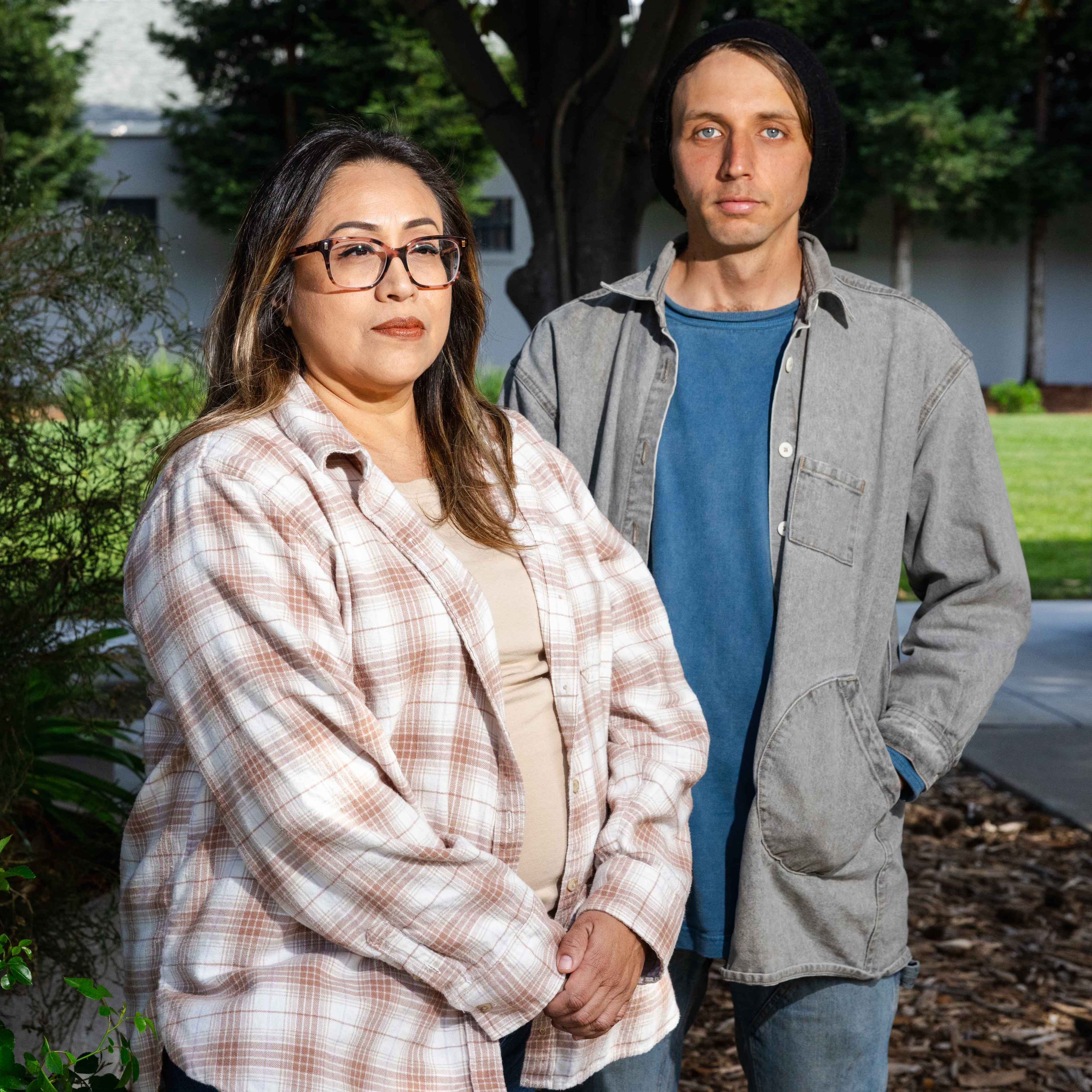 A woman and a man stand outdoors, both looking at the camera. The woman wears glasses and a plaid shirt, while the man has a beanie and a gray overshirt.
