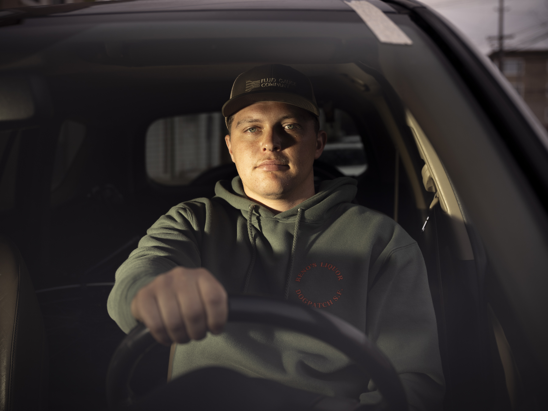 A person is sitting in a car, wearing a cap and a hoodie. They grip the steering wheel with a focused expression, illuminated by soft light.