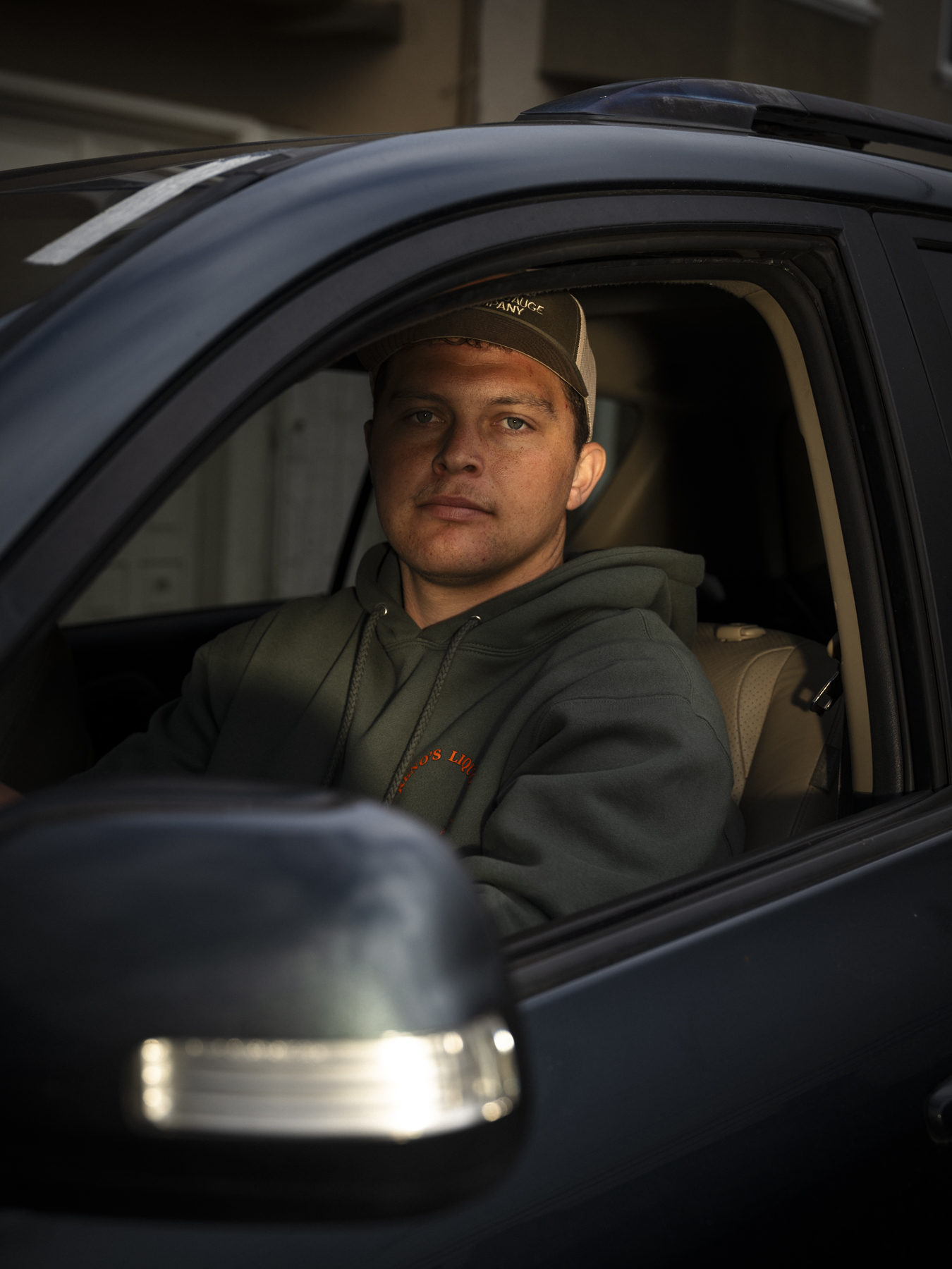 A person sits in the driver's seat of a dark vehicle, wearing a green hoodie and a cap. The scene is dimly lit, emphasizing their focused expression.