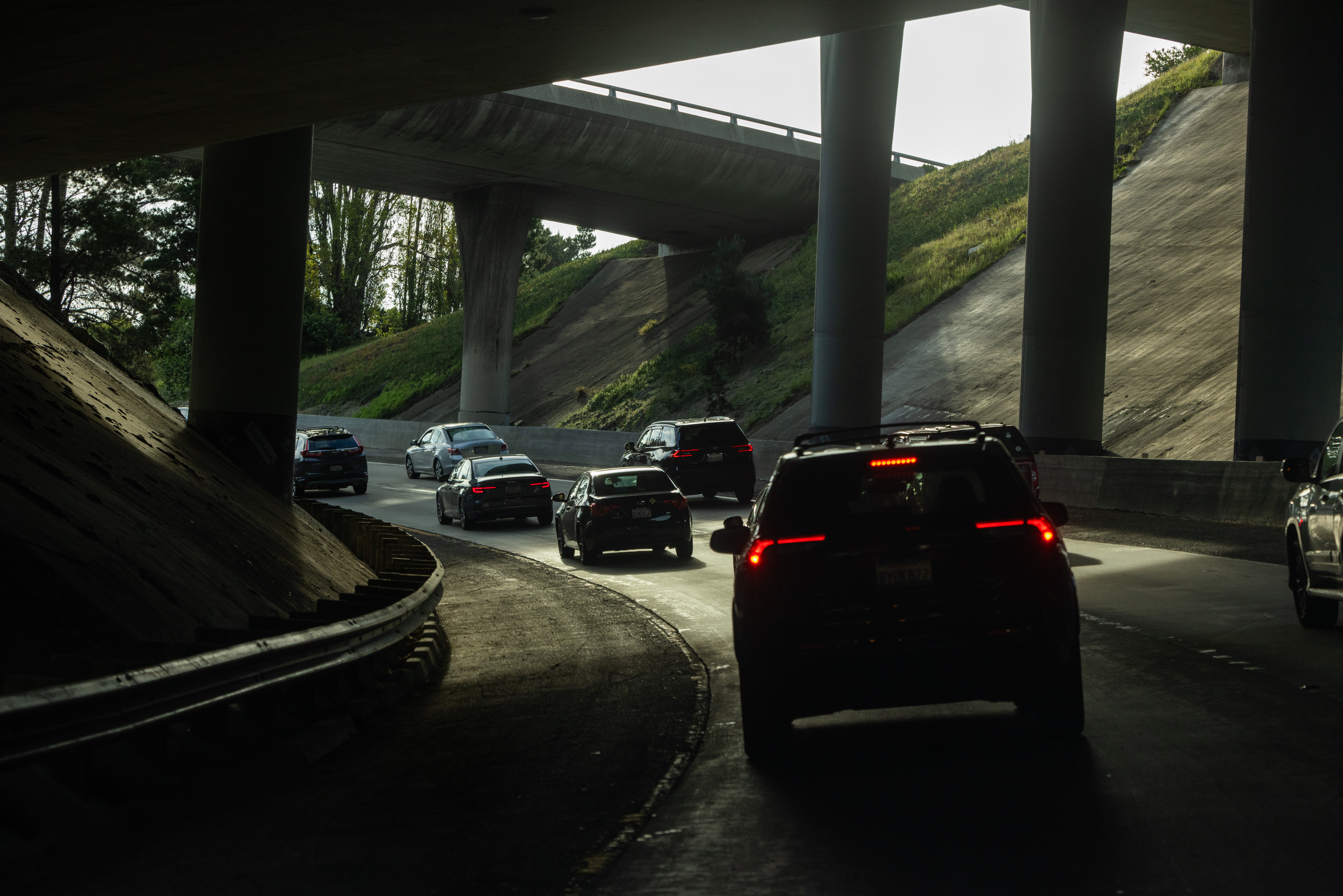 Cars are driving on a shaded road under an overpass, with sunlight filtering through and casting shadows. The road curves, surrounded by greenery.