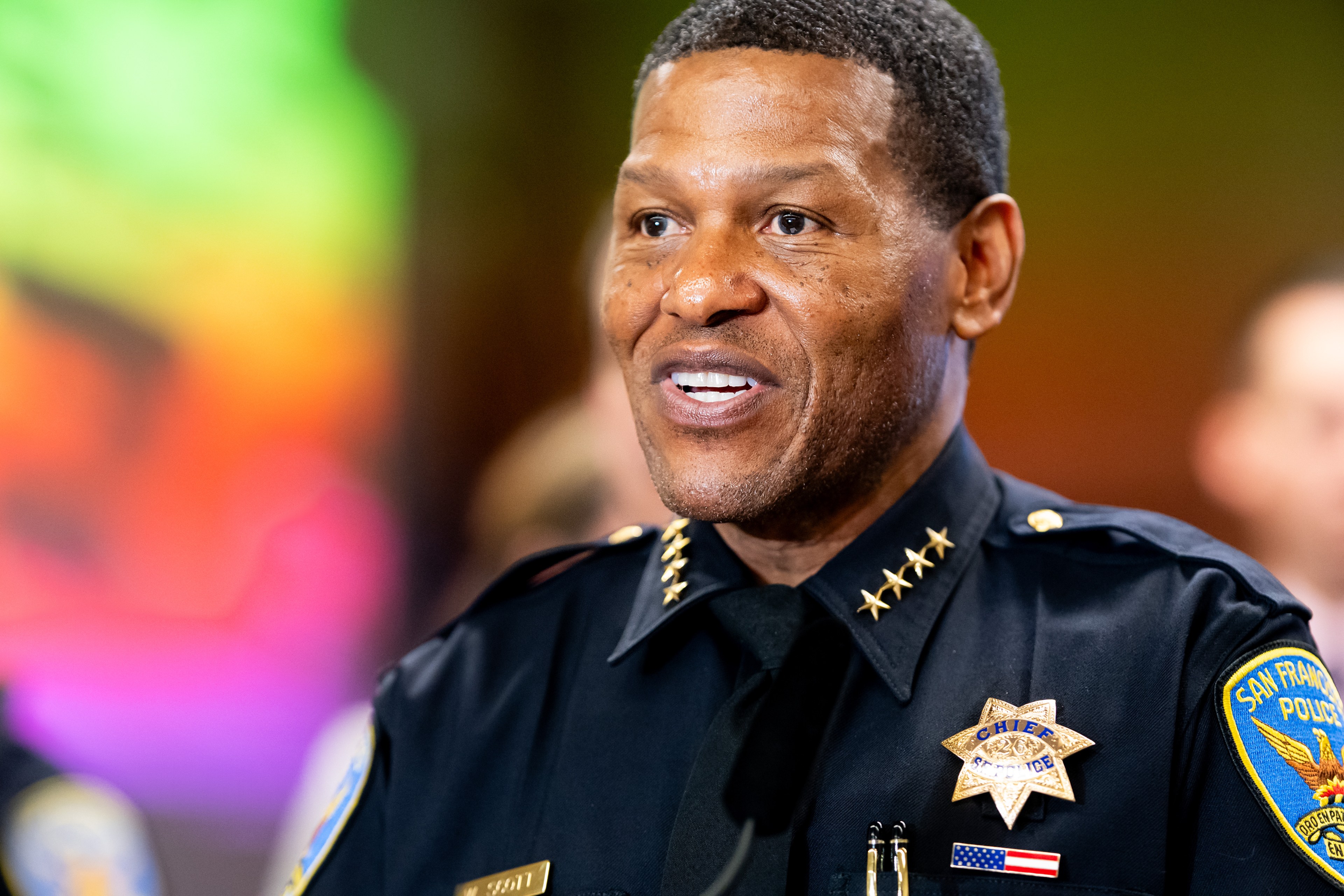 A police officer in uniform is smiling. His uniform displays badges, stars, and an American flag. The background is colorful and blurred.