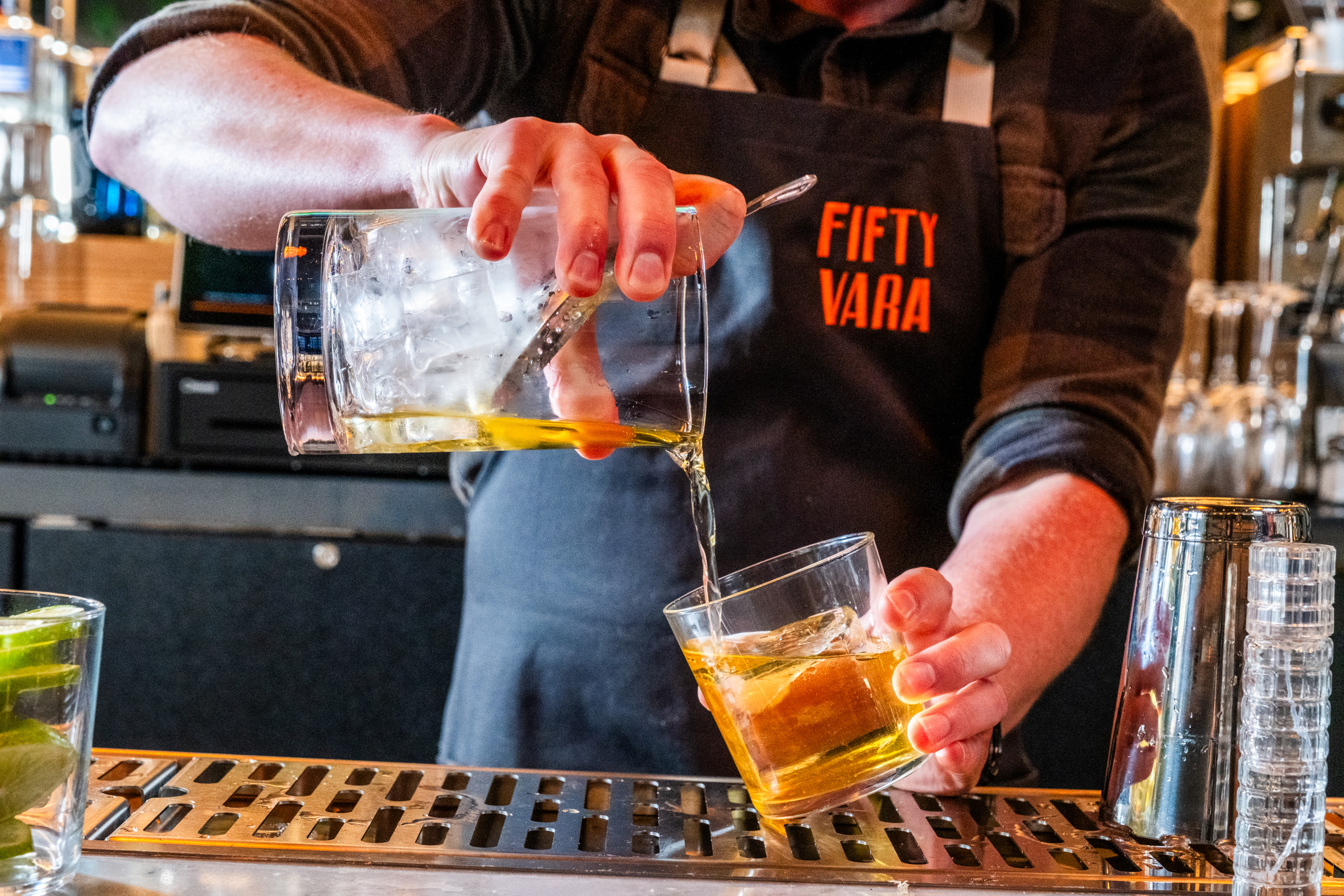 A bartender wearing a &quot;Fifty Vara&quot; apron is pouring a yellowish drink from a mixing glass into a glass with ice on a bar counter.