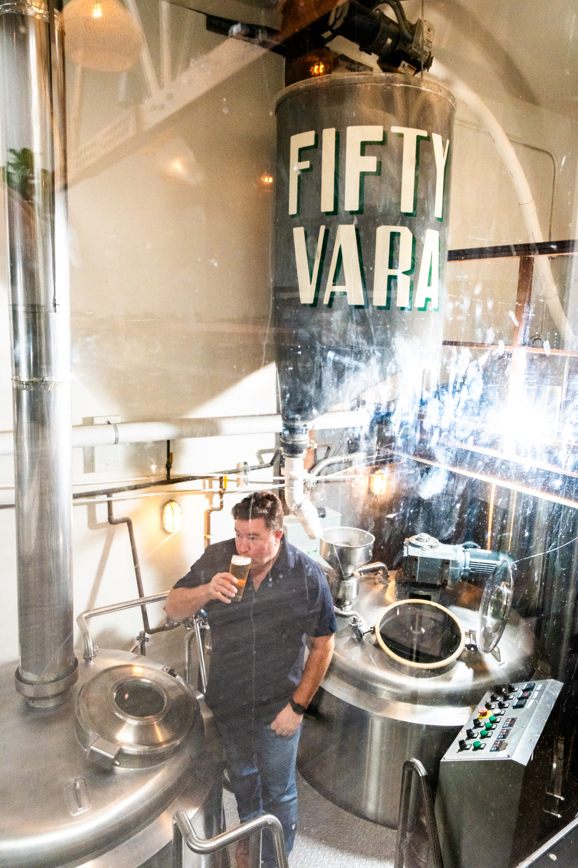 A person stands in a brewery, drinking a beer beside shiny metal brewing equipment. Above is a large container labeled &quot;Fifty Vara.&quot;